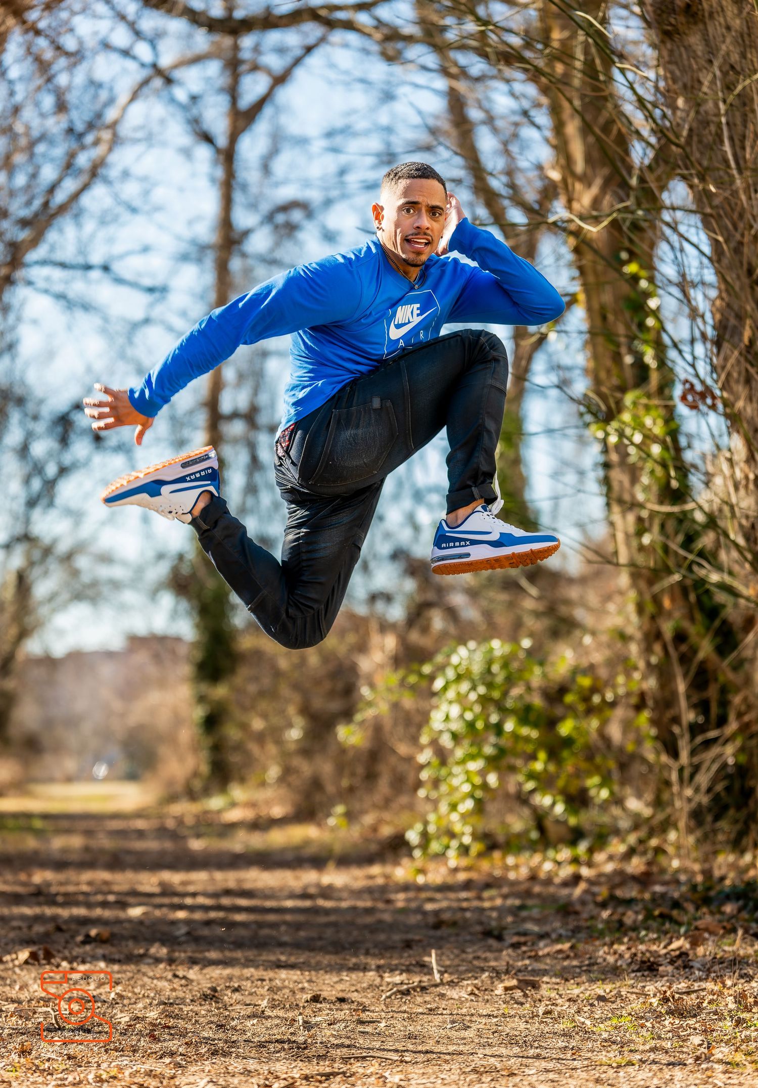 Athletic figure in blue Nike sweatshirt and dark pants performs dynamic jump on dirt trail through autumn woods.