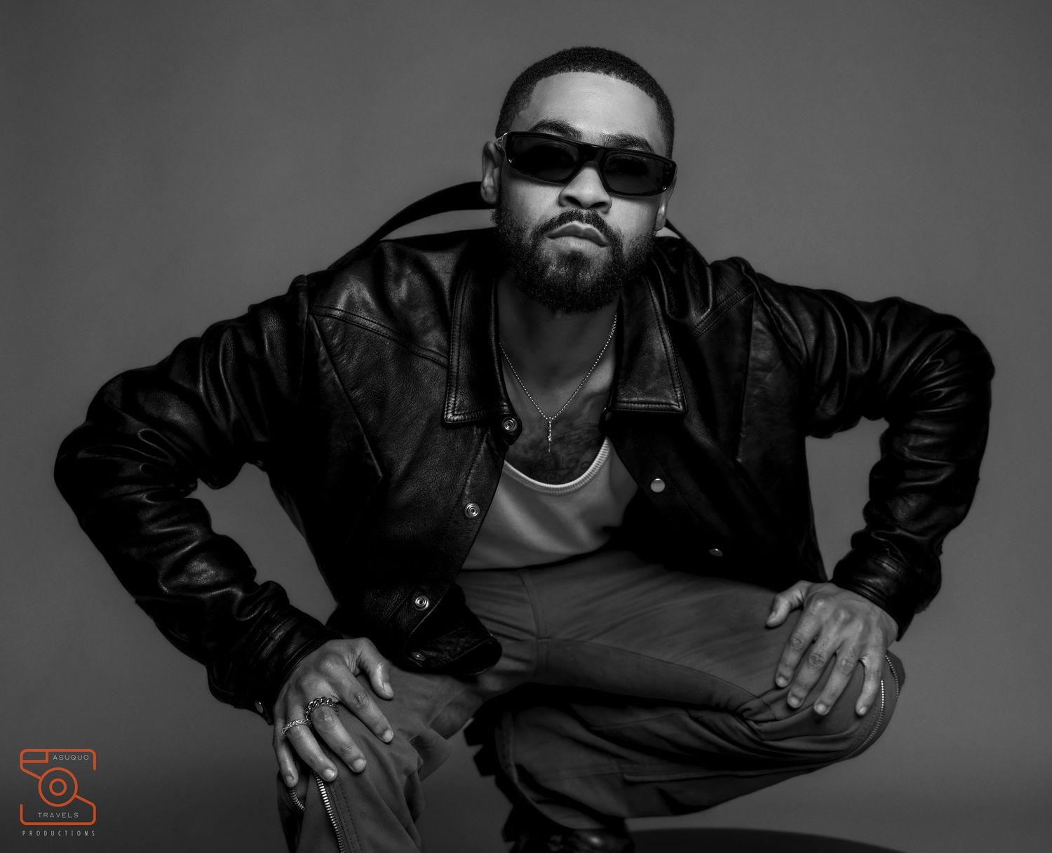 Black and white portrait of a person in leather jacket and sunglasses crouching in a stylized pose.