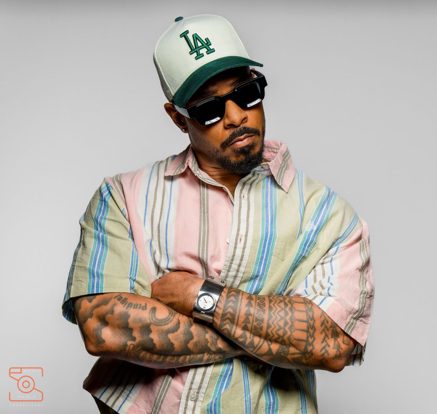 Tattooed man in LA cap, sunglasses, and striped shirt poses with arms crossed in a studio setting.