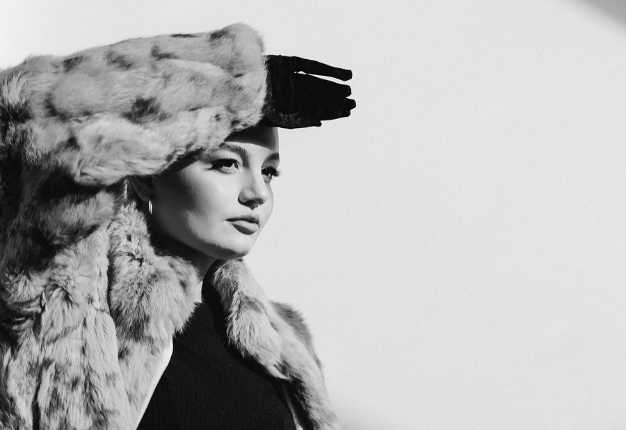 Elegant woman in luxurious fur coat and hat poses in dramatic black-and-white fashion portrait.