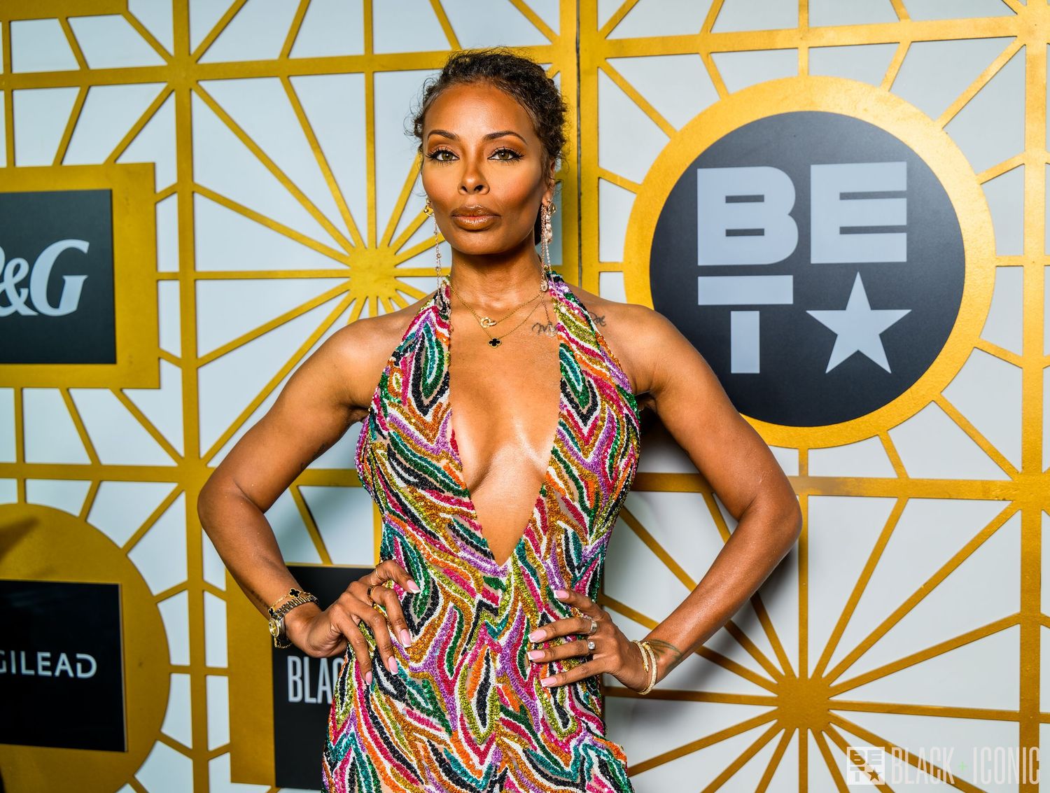 A person poses in a vibrant geometric print dress against a golden art deco patterned backdrop at an event.