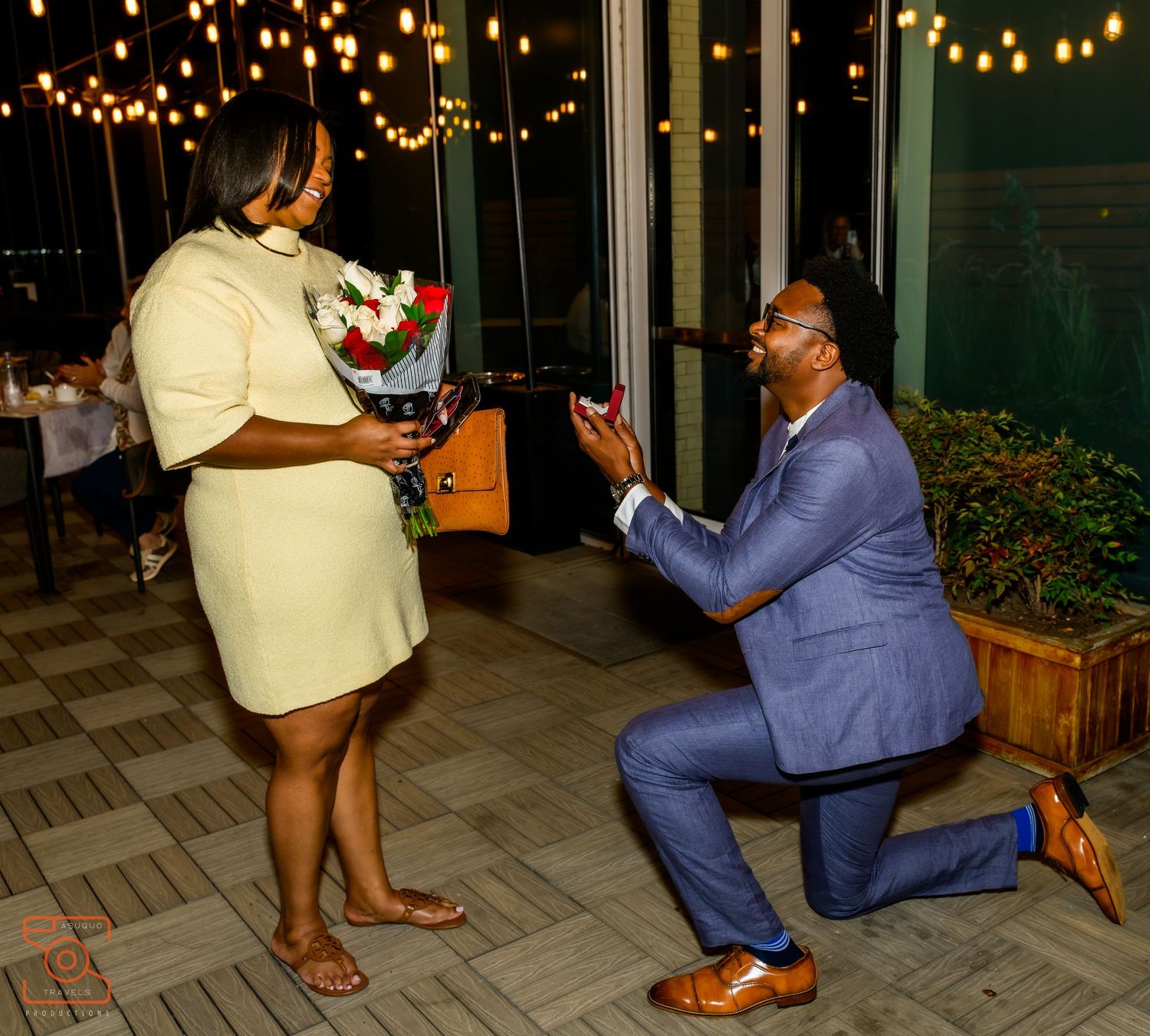 A person in a blue suit kneels down during a romantic outdoor proposal under string lights at night.