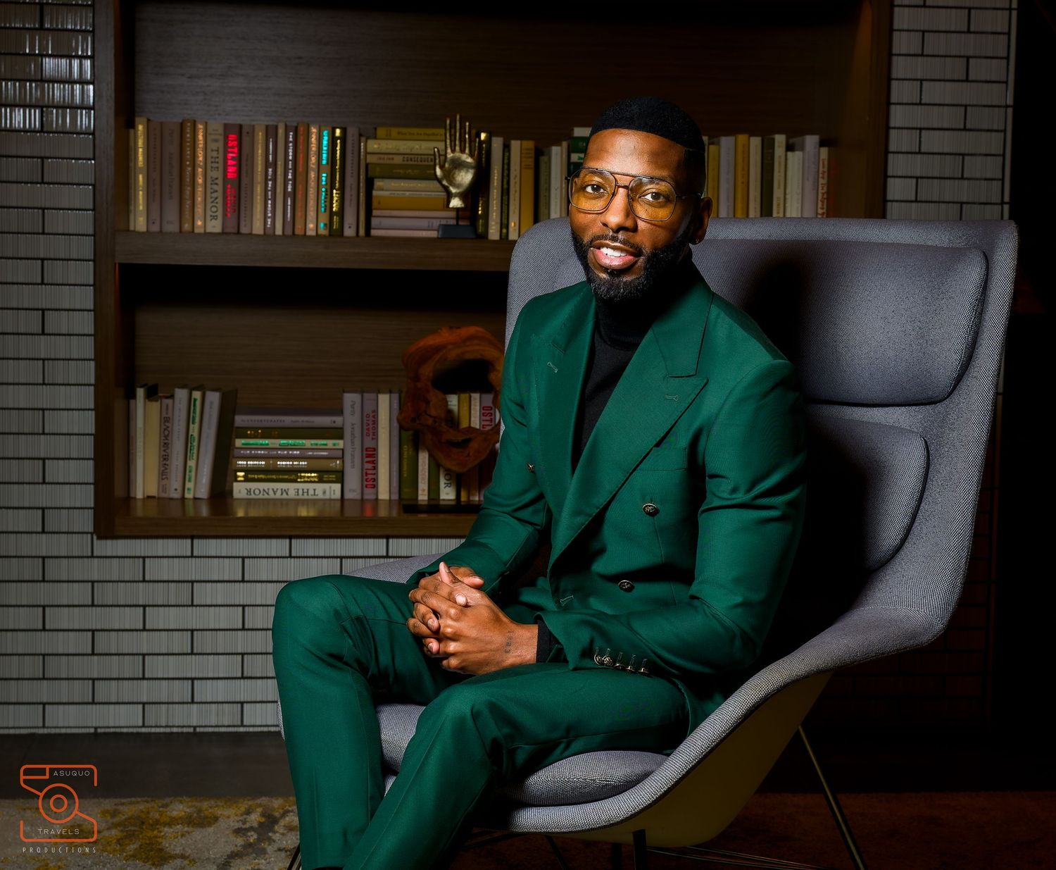 A person wearing an emerald green suit sits in a modern gray chair against a brick wall with bookshelves.