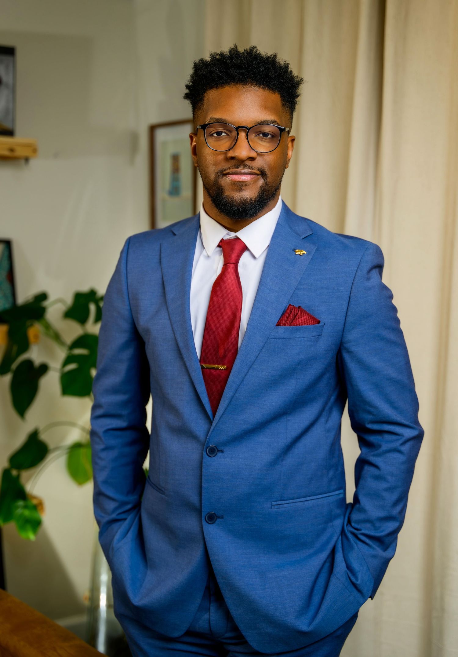 A professional man in a blue suit with red tie and pocket square, wearing glasses, posing confidently indoors.