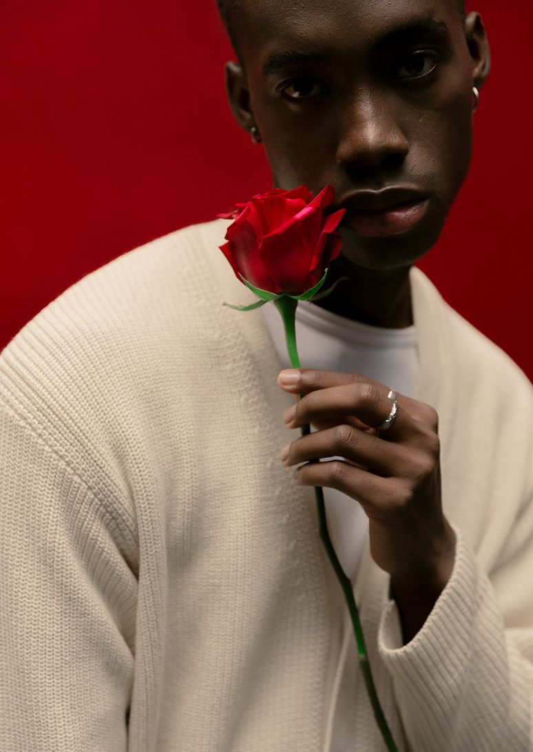 Young Black man in cream sweater holds a red rose near his face against a bold red background.