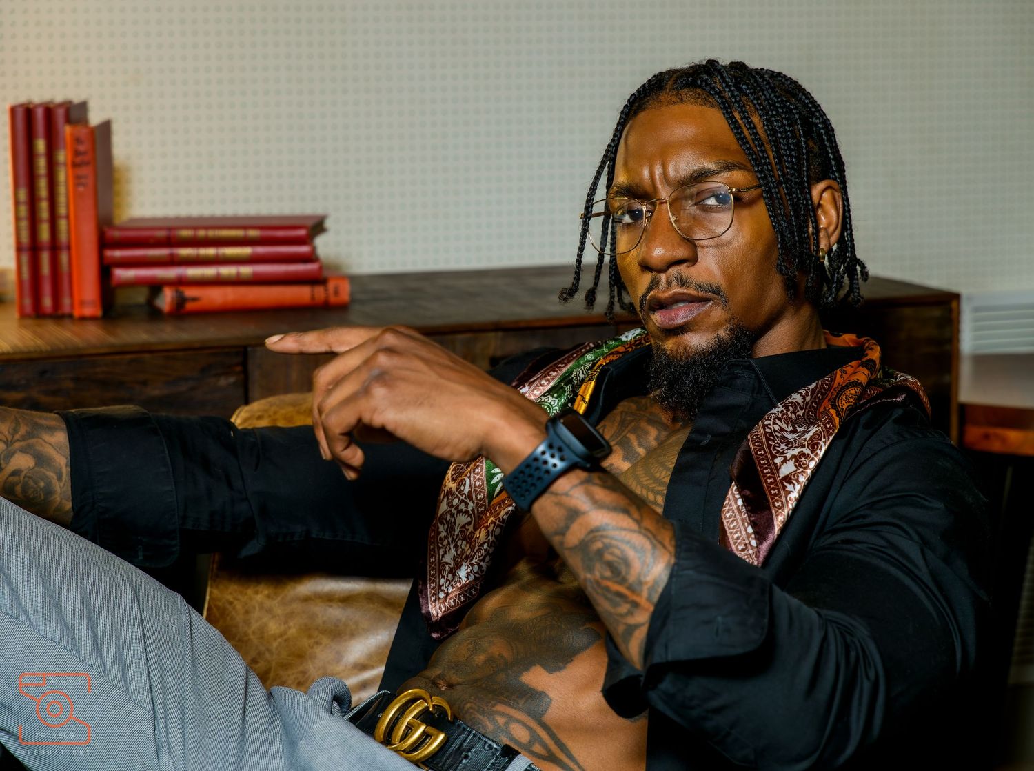 A person with braided hair and tattoos reclines on a couch wearing a black shirt and gold jewelry.