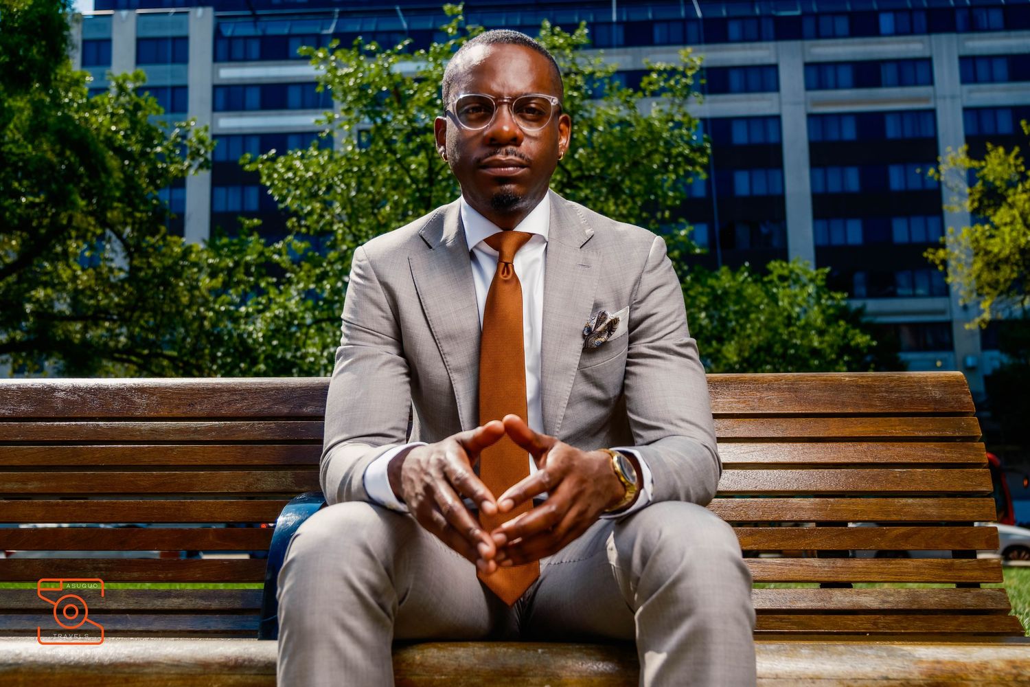 Professional in a light gray suit and orange tie sits confidently on a wooden bench with modern buildings in background.