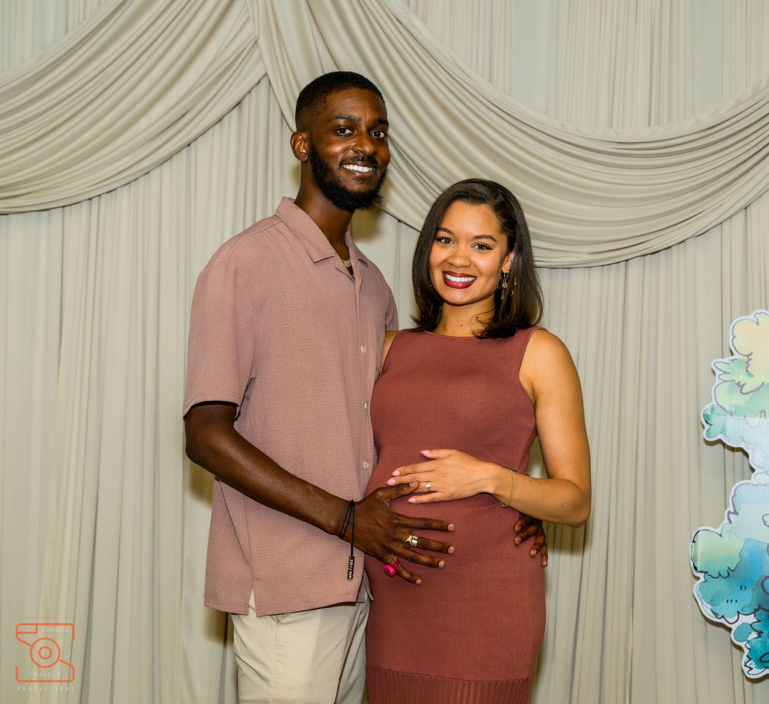 Smiling couple poses together at a baby shower, woman in burgundy dress cradling baby bump, man in mauve shirt beside her.