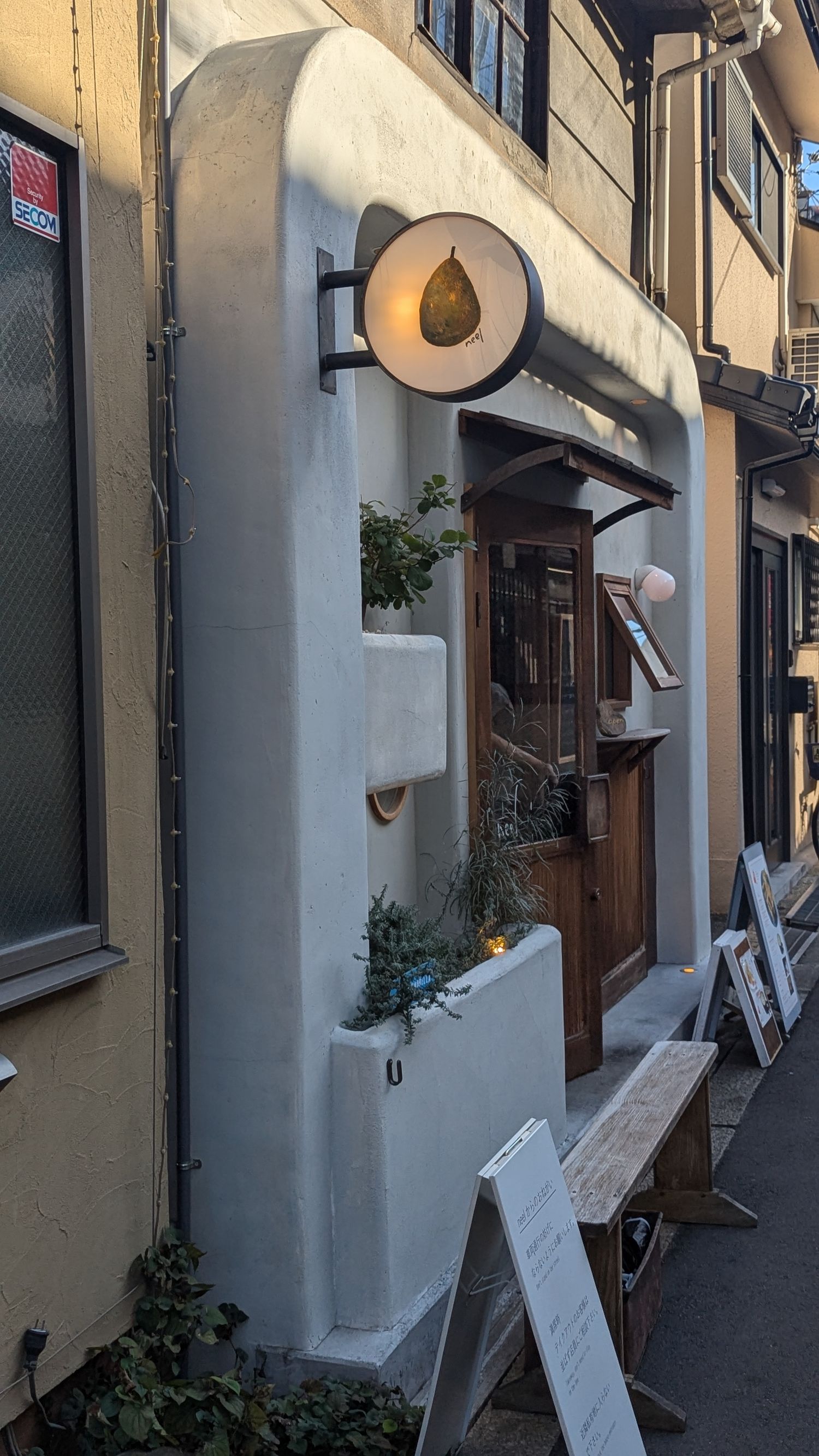 A cozy storefront entrance with a white facade, illuminated pear-shaped sign, and small decorative plants along the wall.