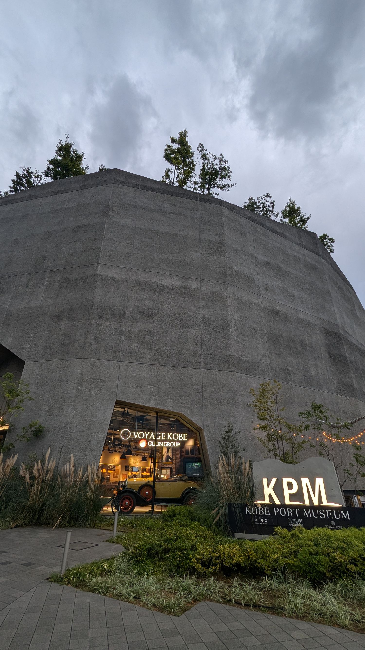 Modern museum entrance with concrete curved wall and KPM signage under dramatic cloudy sky in Osaka.
