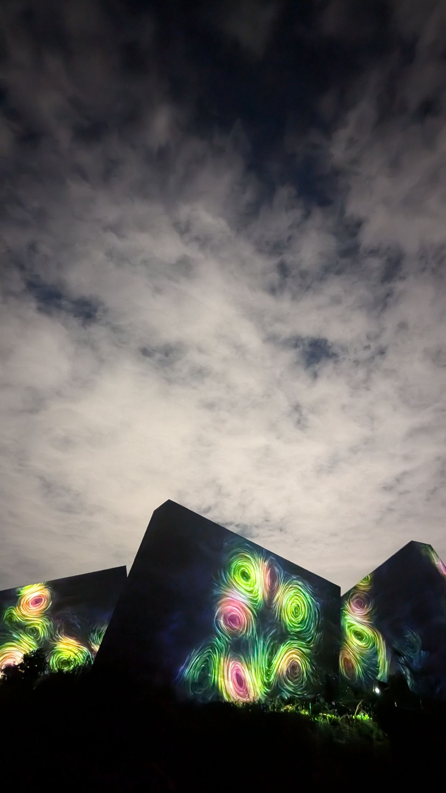 Glowing neon peacock patterns illuminate a dark silhouetted mountain ridge against a dramatic cloudy night sky.