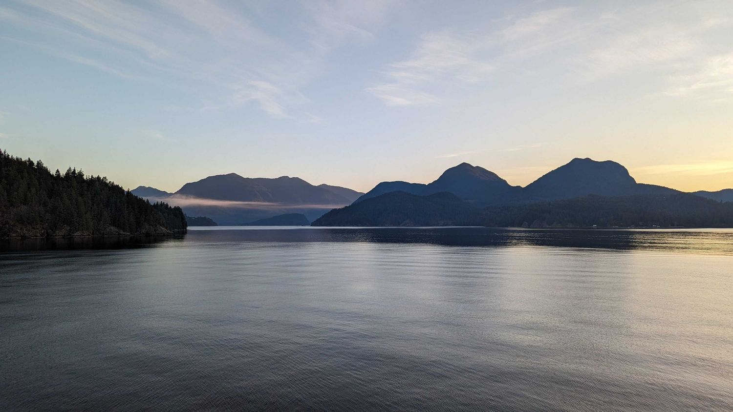 A tranquil panoramic view of a calm lake surrounded by mountains and evergreen forests during sunset.