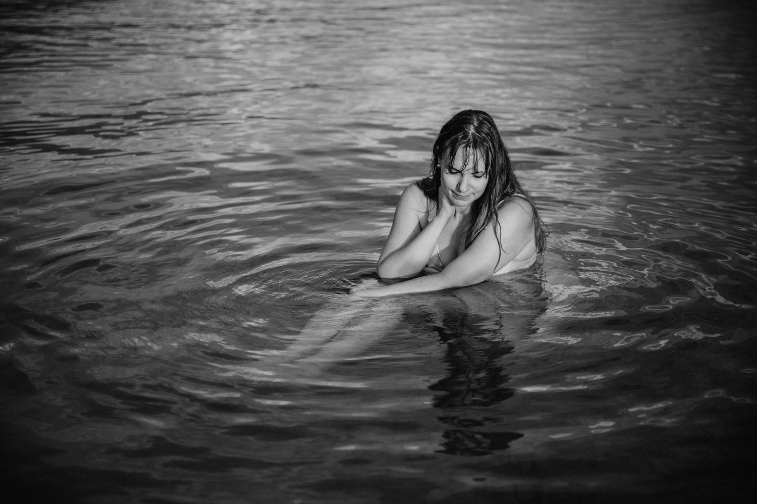 Boudoir-Foto in Schwarz-Weiss: Frau in nassem Kleid umarmt ihre Beine und sitzt still im Wasser – sinnlich, verletzlich, ruhig.