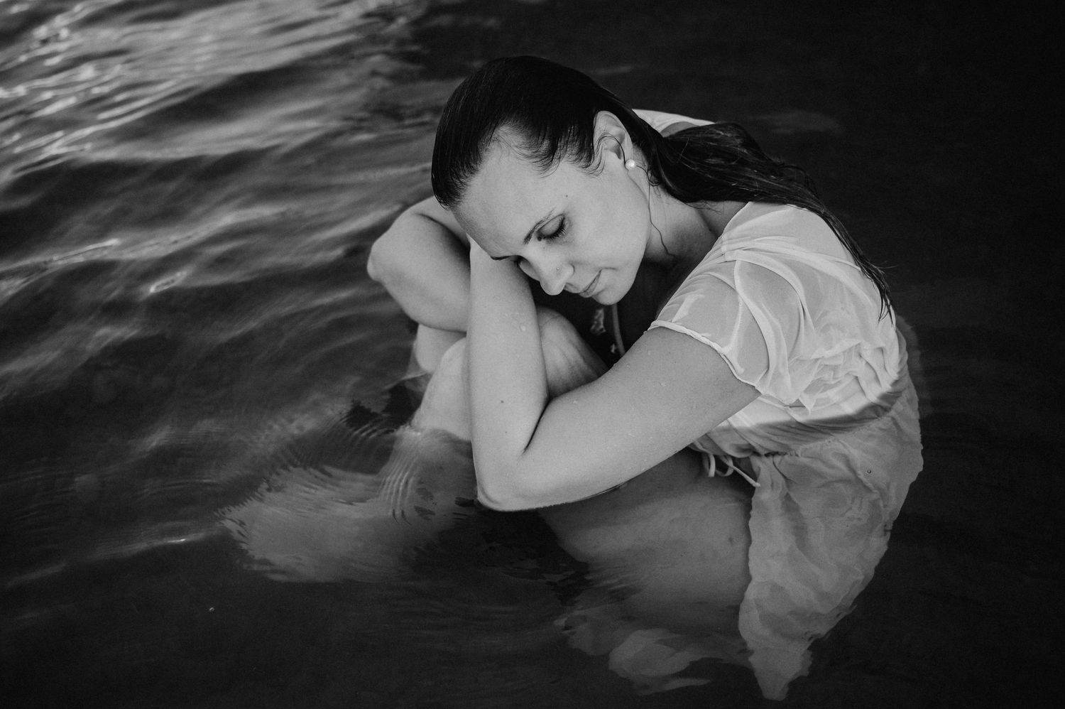 Boudoir-Foto in Schwarz-Weiss: Frau in nassem Kleid umarmt ihre Beine und sitzt still im Wasser – sinnlich, verletzlich, ruhig.