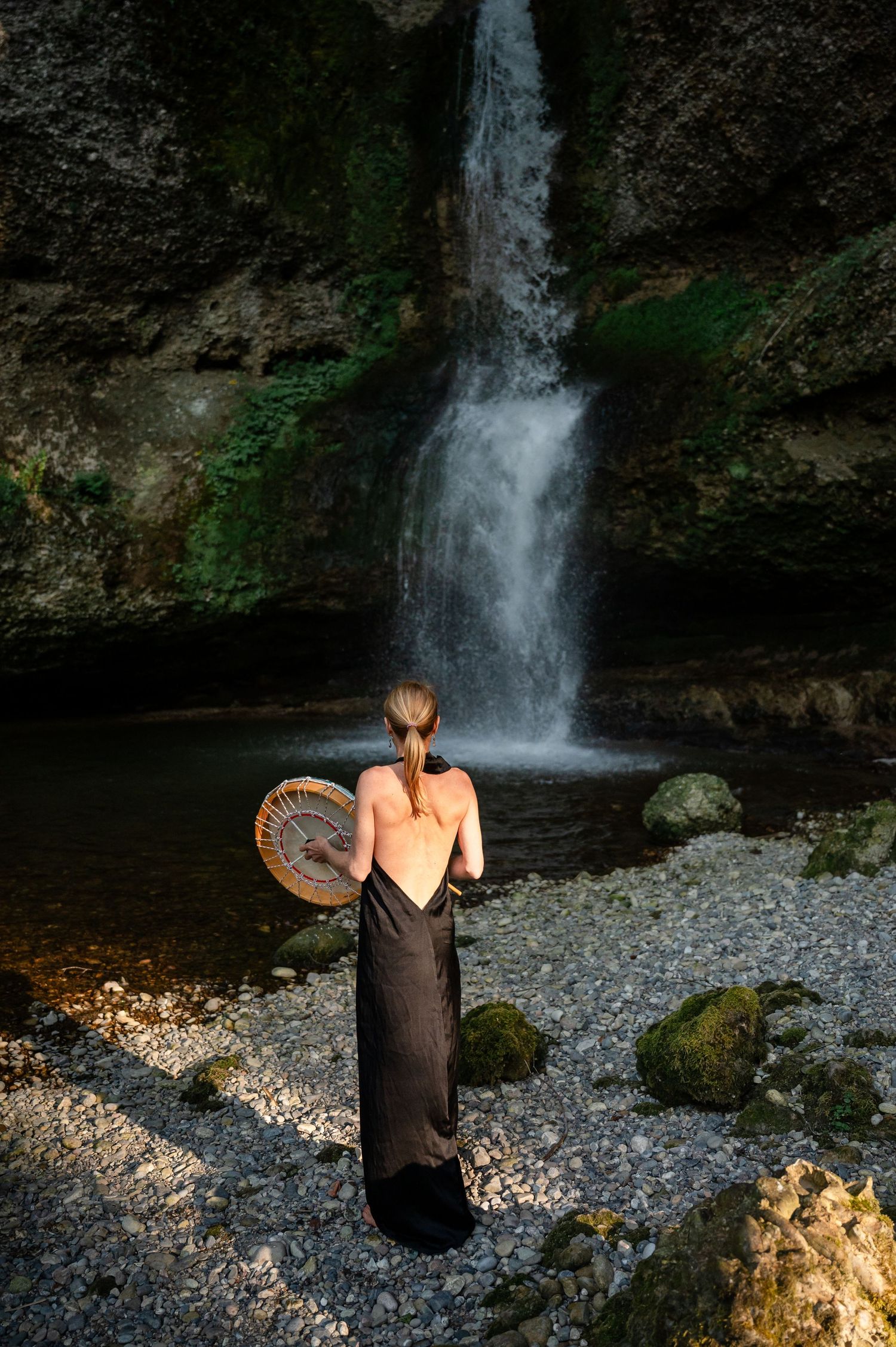 Frau mit schamanischer Trommel steht im langen Kleid vor Wasserfall – spirituelle Verbindung zur Natur im Abendlicht.