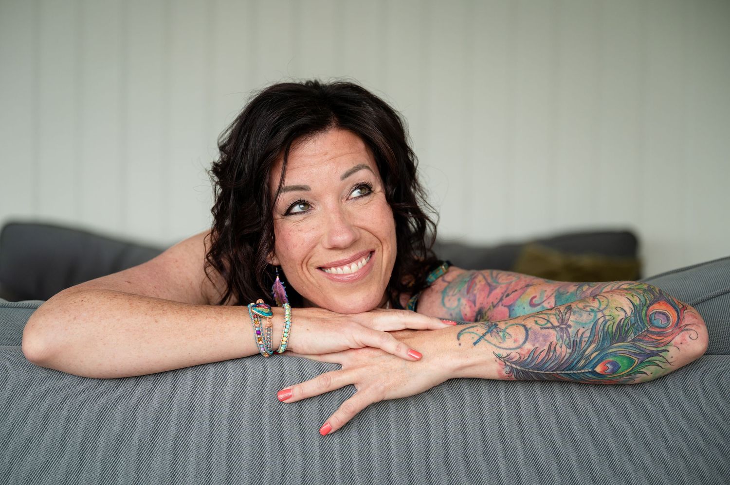 Fröhliche Frau mit buntem Tattoo lehnt entspannt über die Sofalehne und strahlt – authentisches Selbstliebe-Shooting in Thun.