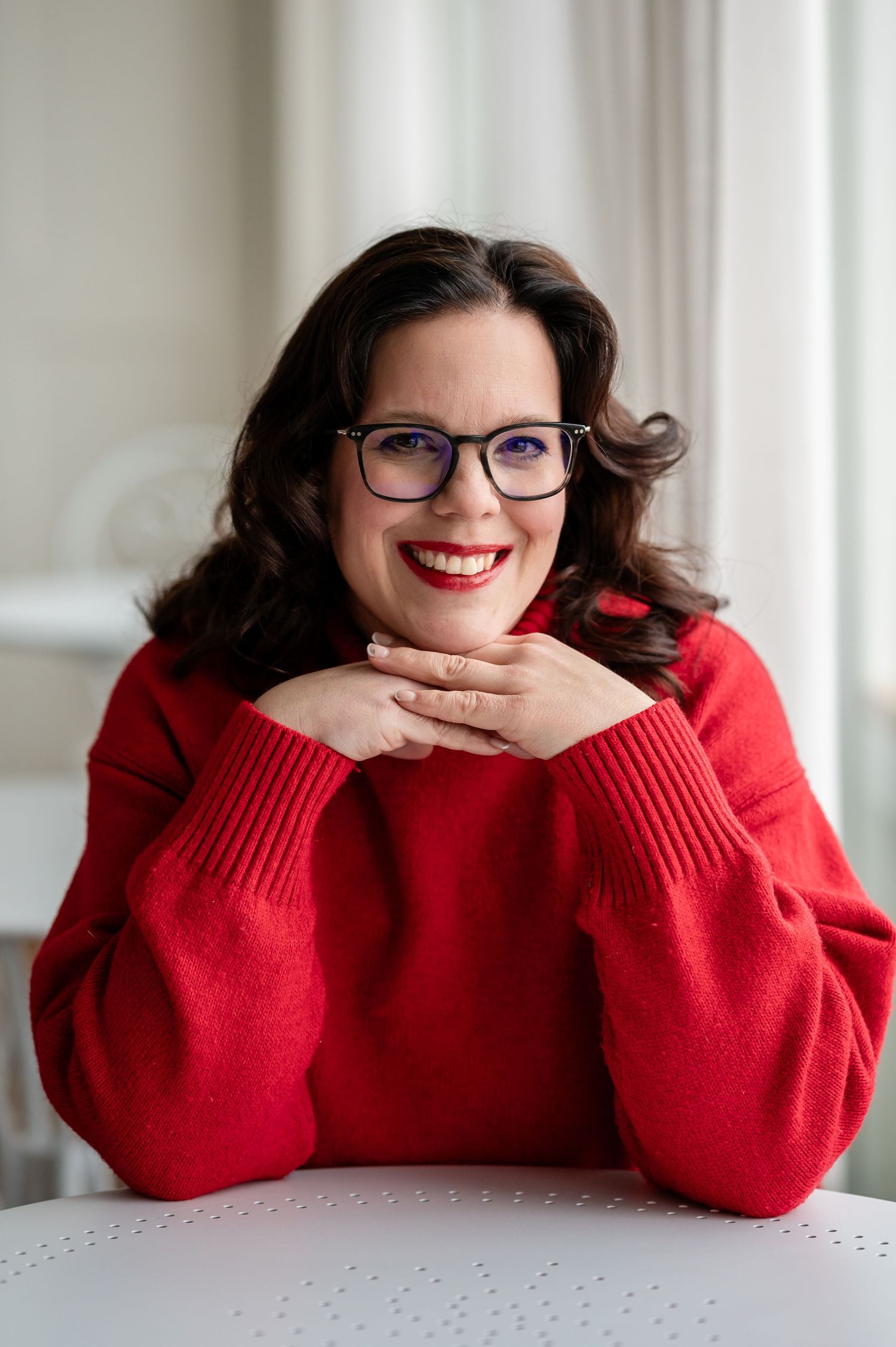 Frau mit Brille und rotem Pullover lächelt strahlend in die Kamera, stützt das Kinn auf die Hände – Selbstliebe-Shooting in Thun.