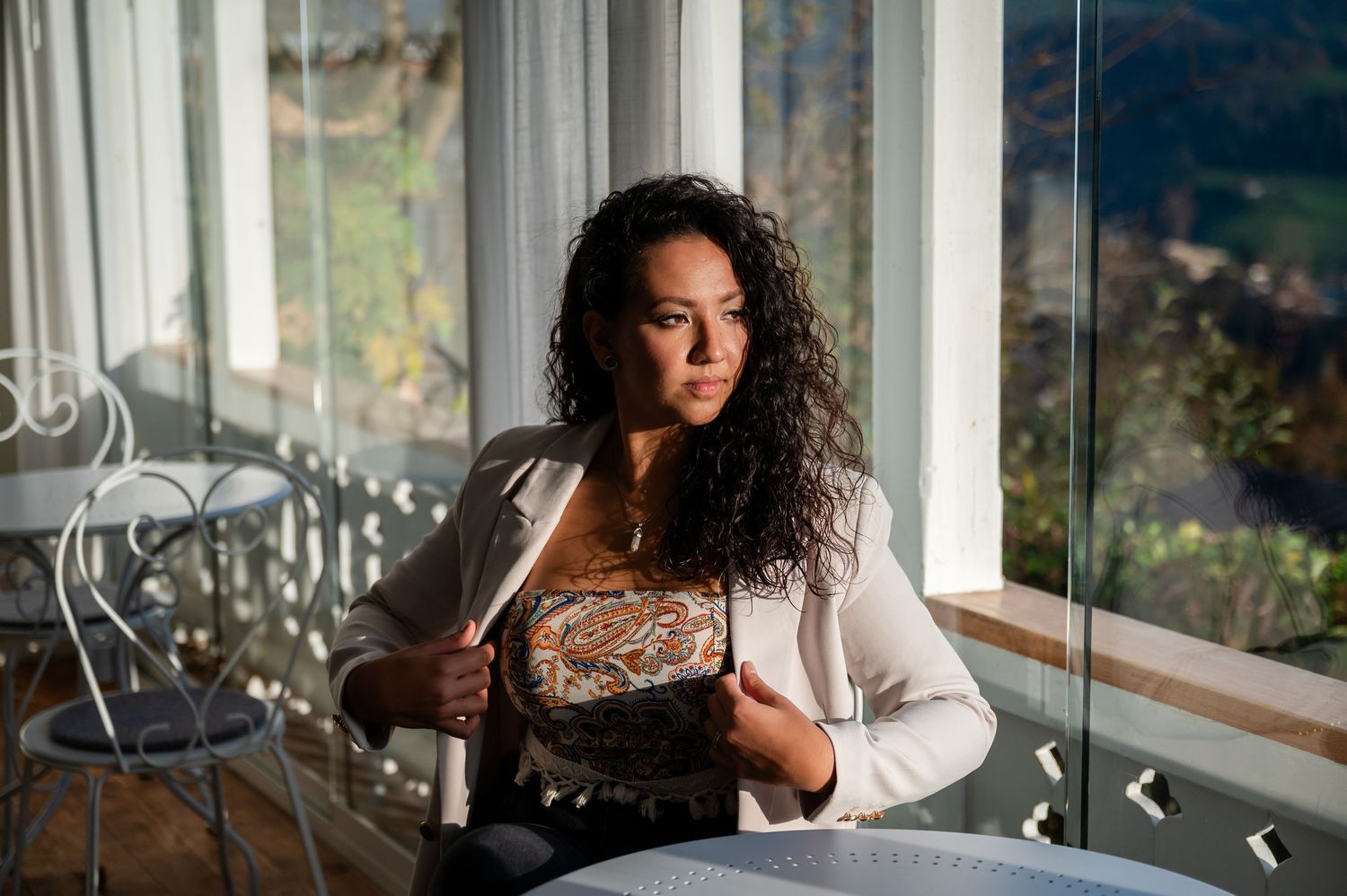 Selbstbewusste Frau mit lockigem Haar sitzt im Sonnenlicht am Fenster, Blick nach aussen gerichtet – stimmungsvoll und stark.
