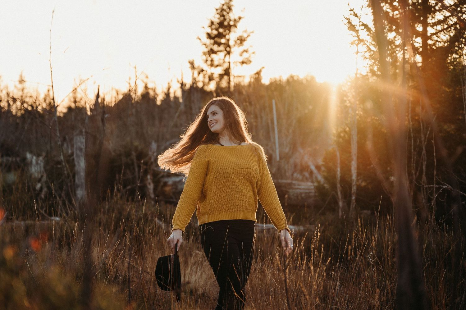 Frau im goldenen Licht der Abendsonne, mit gelbem Pullover und Hut in der Hand – stimmungsvolles Outdoor-Porträt in der Natur.