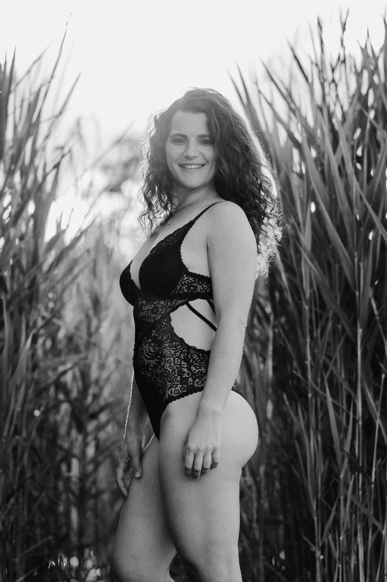 Selbstbewusste Frau in schwarzer Spitze, lächelnd im Schilf am Thunersee – sinnliches Boudoirfoto in Schwarz-Weiss.