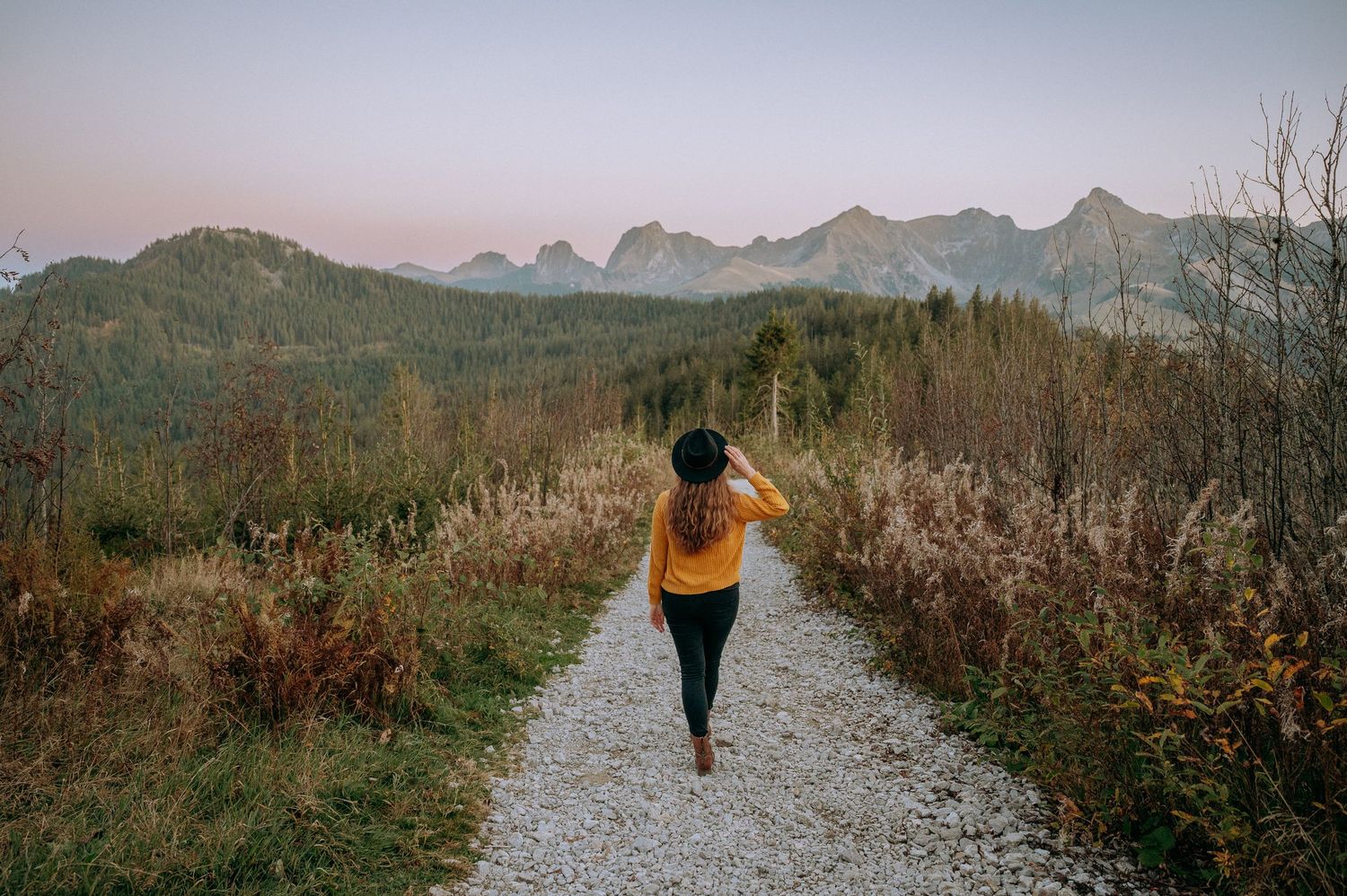 Frau mit Hut spaziert auf Kiesweg im Gantrischgebiet, umgeben von herbstlicher Natur und Bergpanorama – natürliches Outdoor-Porträt.