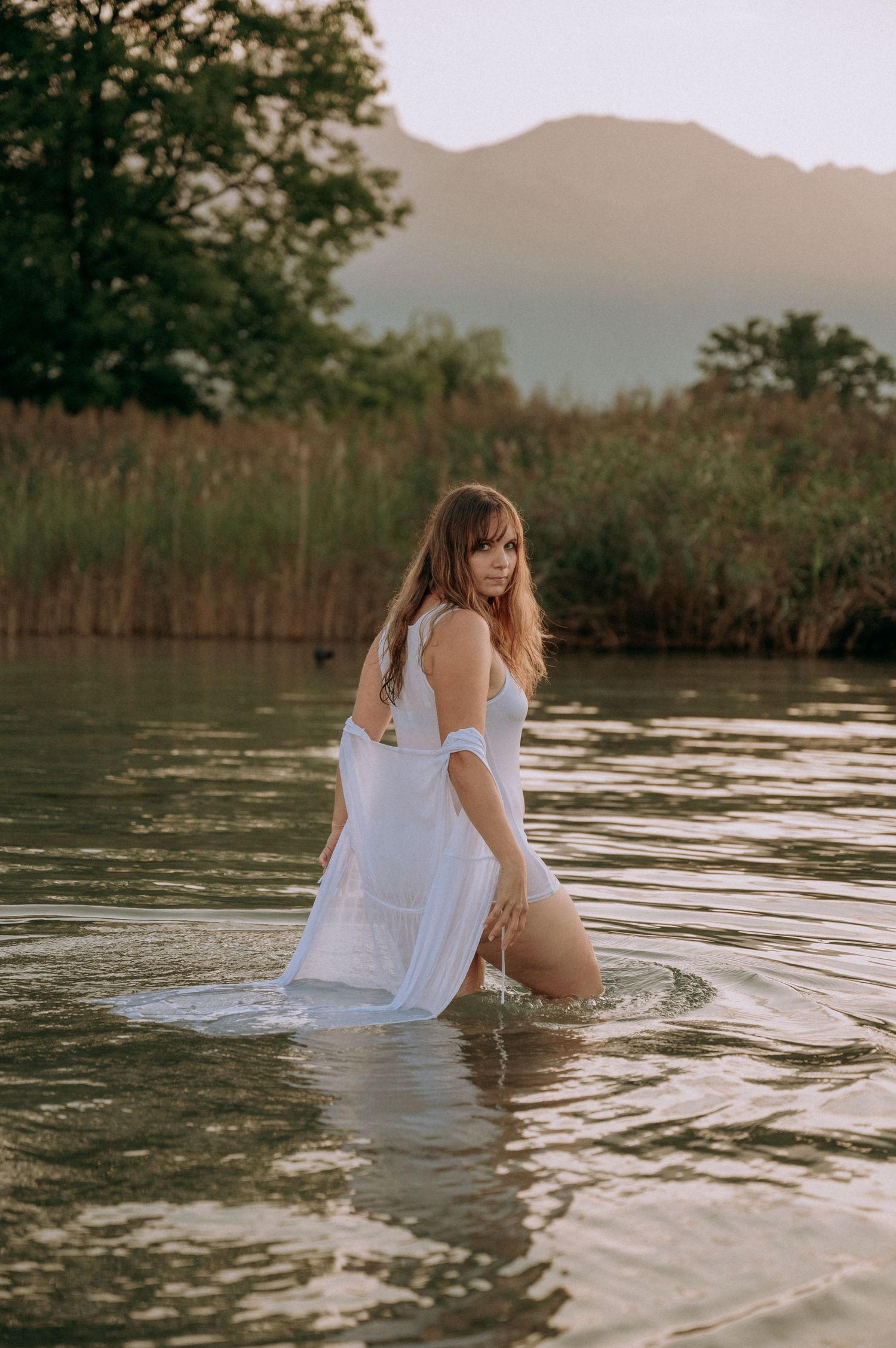 Boudoir-Shooting im See bei Sonnenuntergang: Frau in nassem, weißen Kleid watet sinnlich durchs Wasser – natürlich, ruhig, feminin.