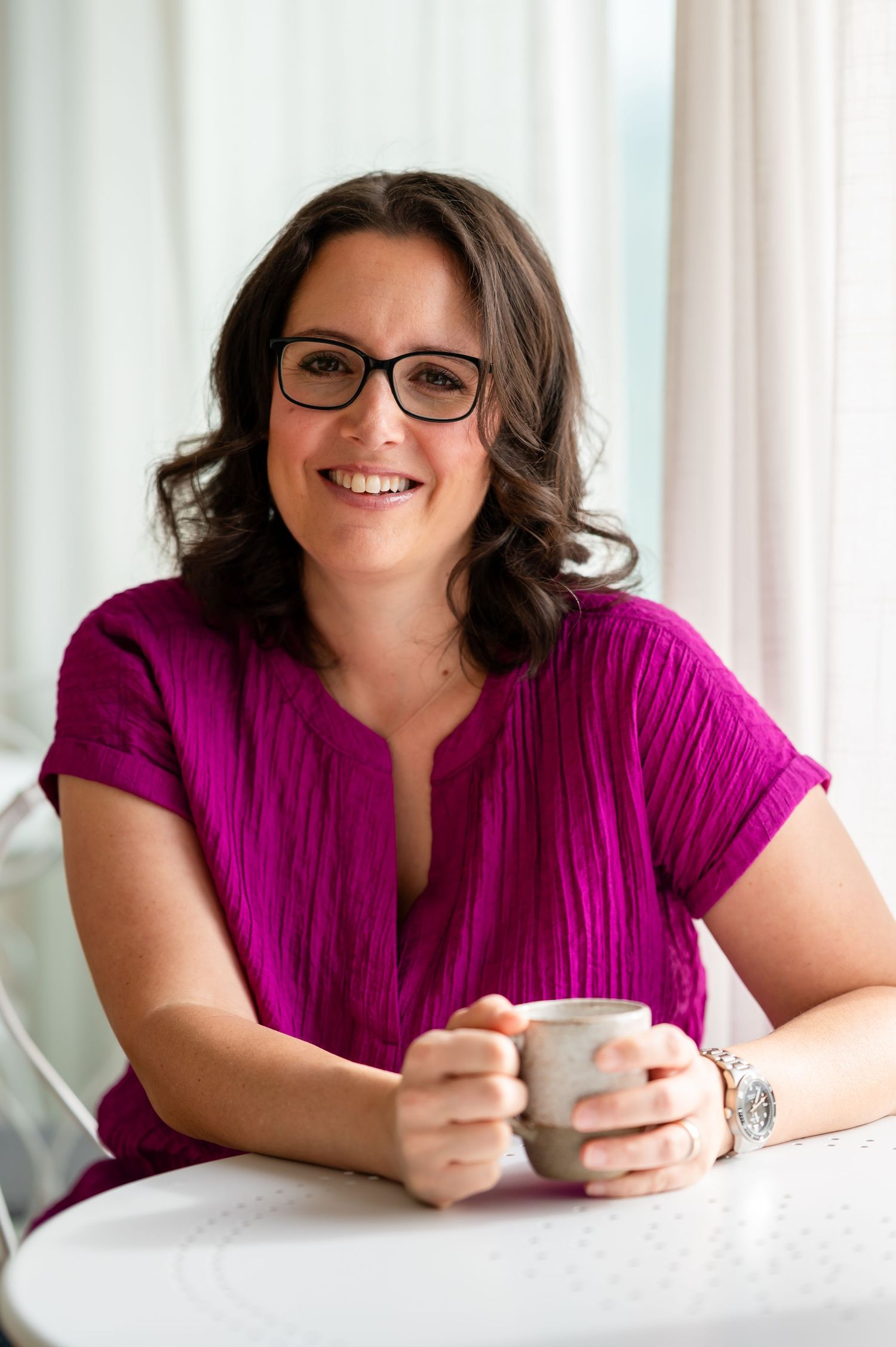 Frau mit lockigem Haar und Brille sitzt lächelnd am Tisch mit einer Tasse in der Hand, in einer hellen Coaching-Atmosphäre in Thun.