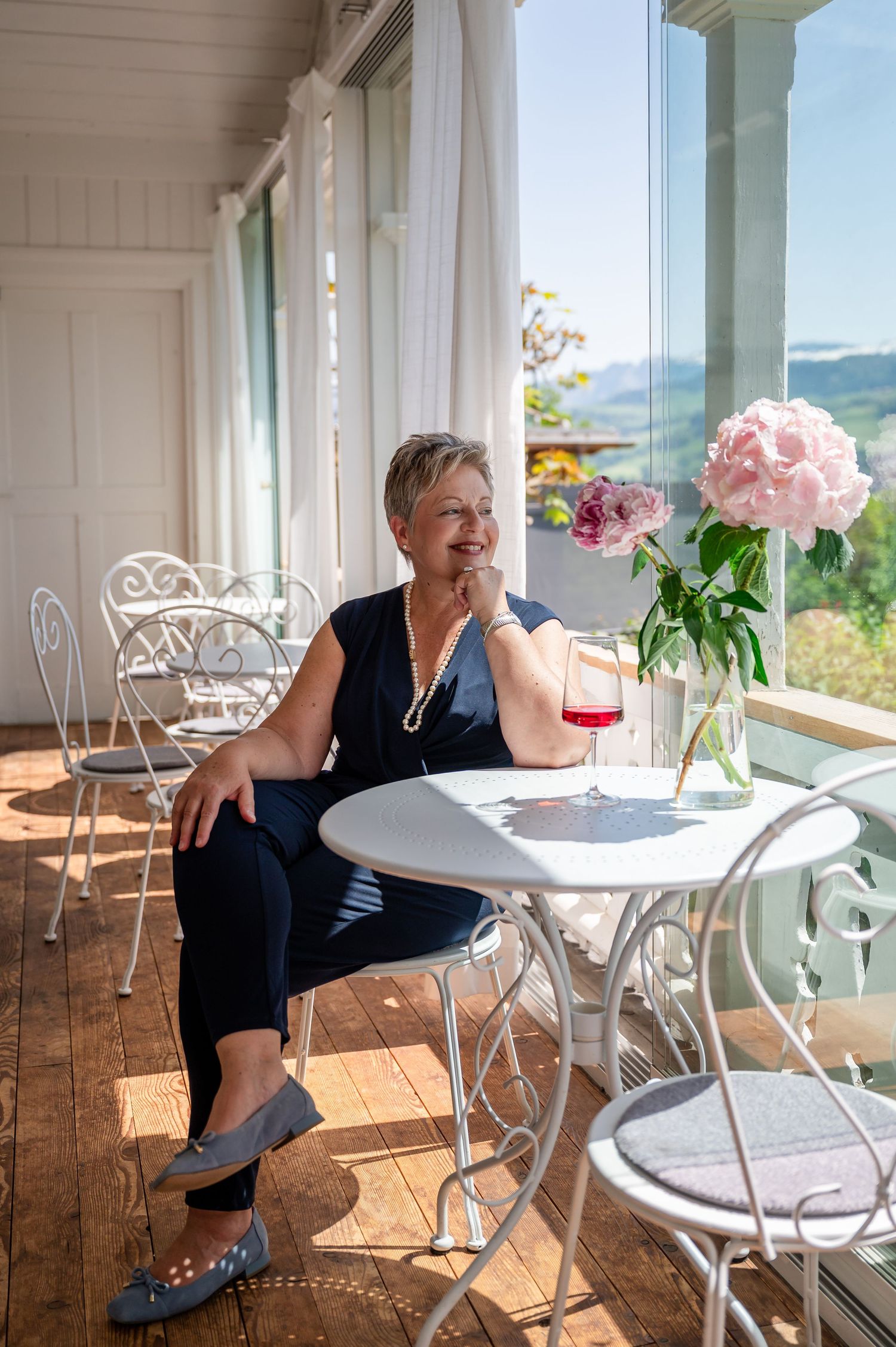 Ältere Frau sitzt entspannt in stilvollem Café mit Blick ins Grüne, genießt den Moment bei einem Glas Rosé – Selbstliebe-Shooting Thun