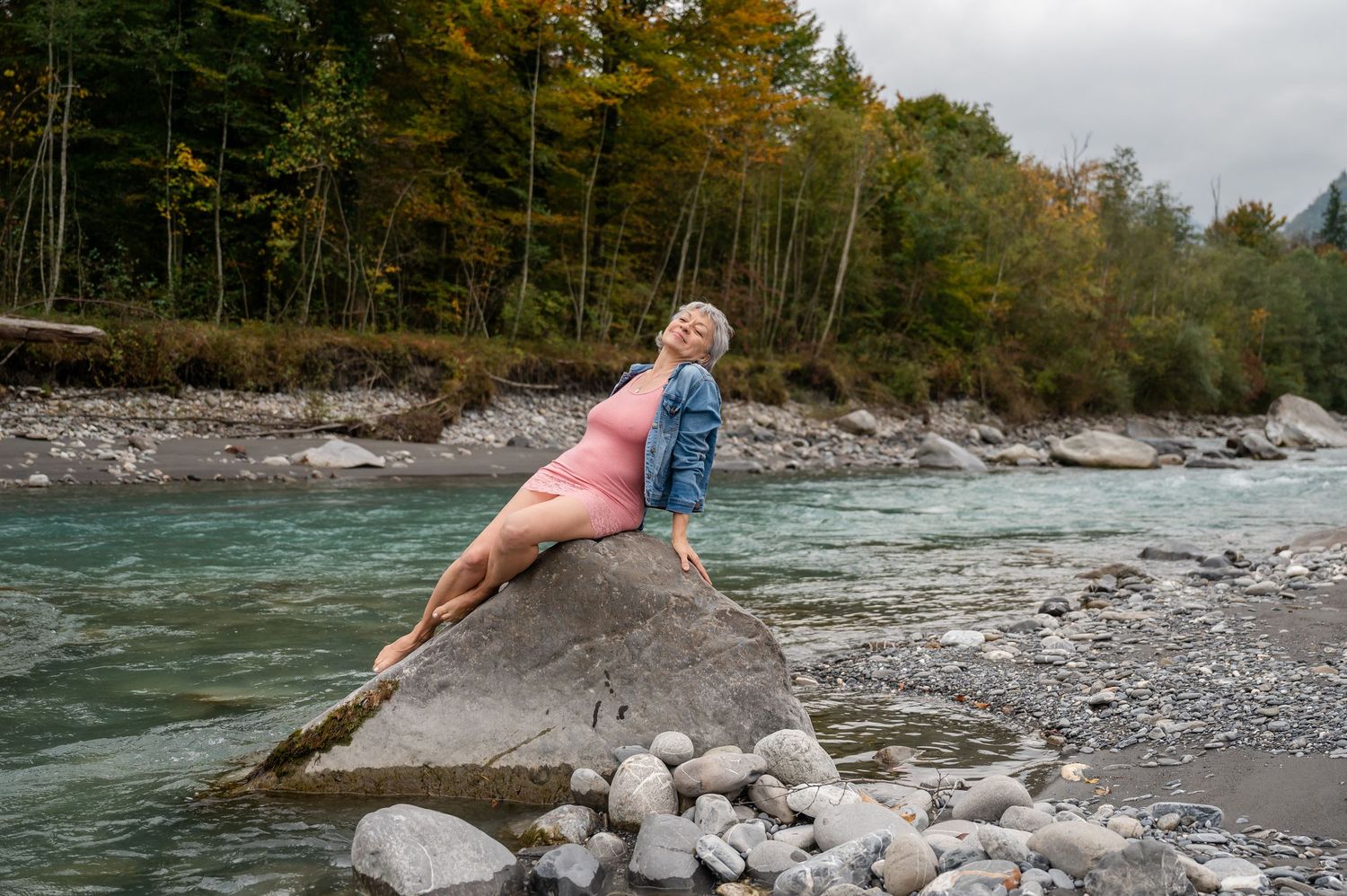 Boudoir-Shooting in der Natur: Frau in rosa Kleid posiert sinnlich an Felsblock am Fluss, Berge im Hintergrund, kraftvoll und wild.