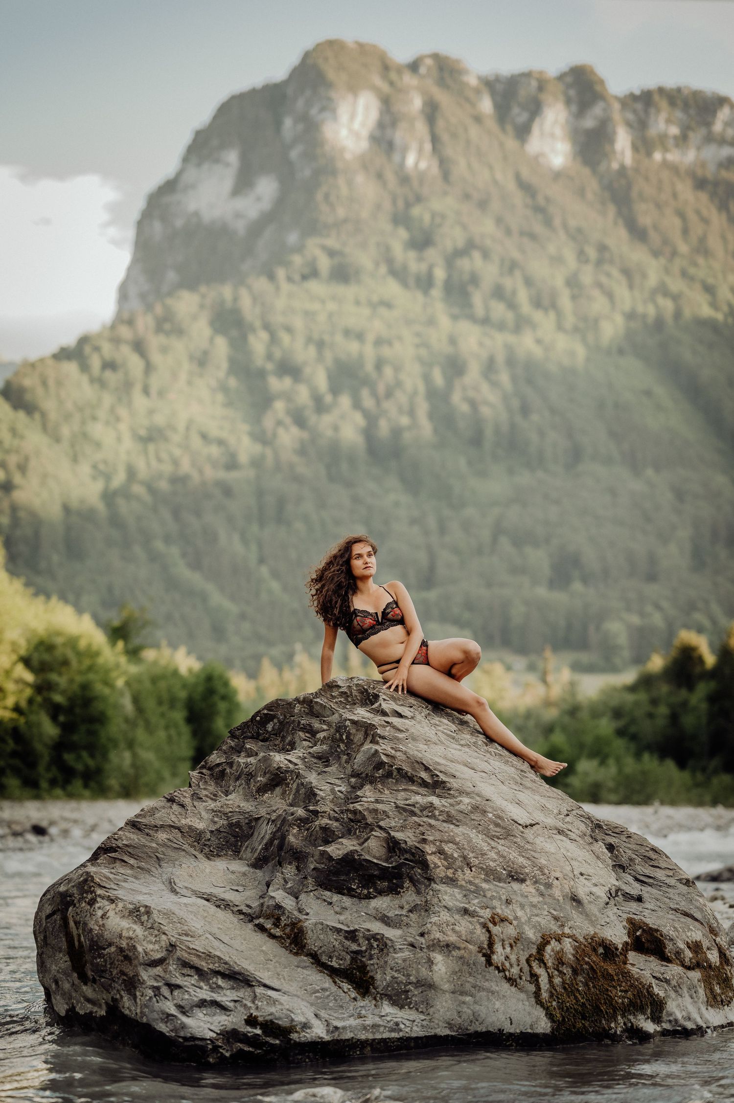 Frau in schwarzer Spitze posiert selbstbewusst auf Fels im Fluss. Sinnliches Boudoirshooting in der Natur bei Thun, Schweiz.