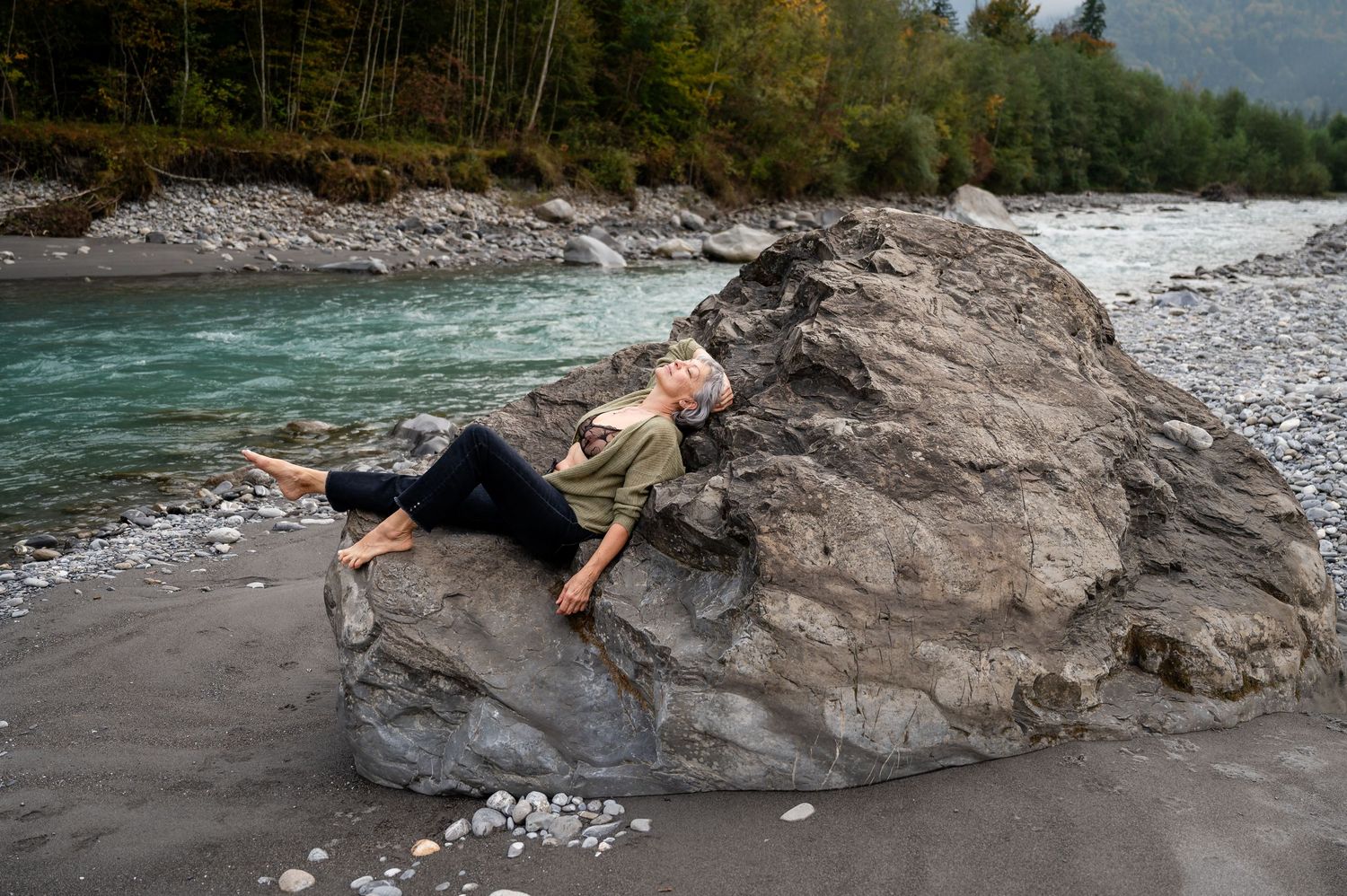 Ältere Frau beim Boudoir-Shooting am Fluss, entspannt und sinnlich in einem grünen Pullover auf Felsen liegend, selbstbewusst.