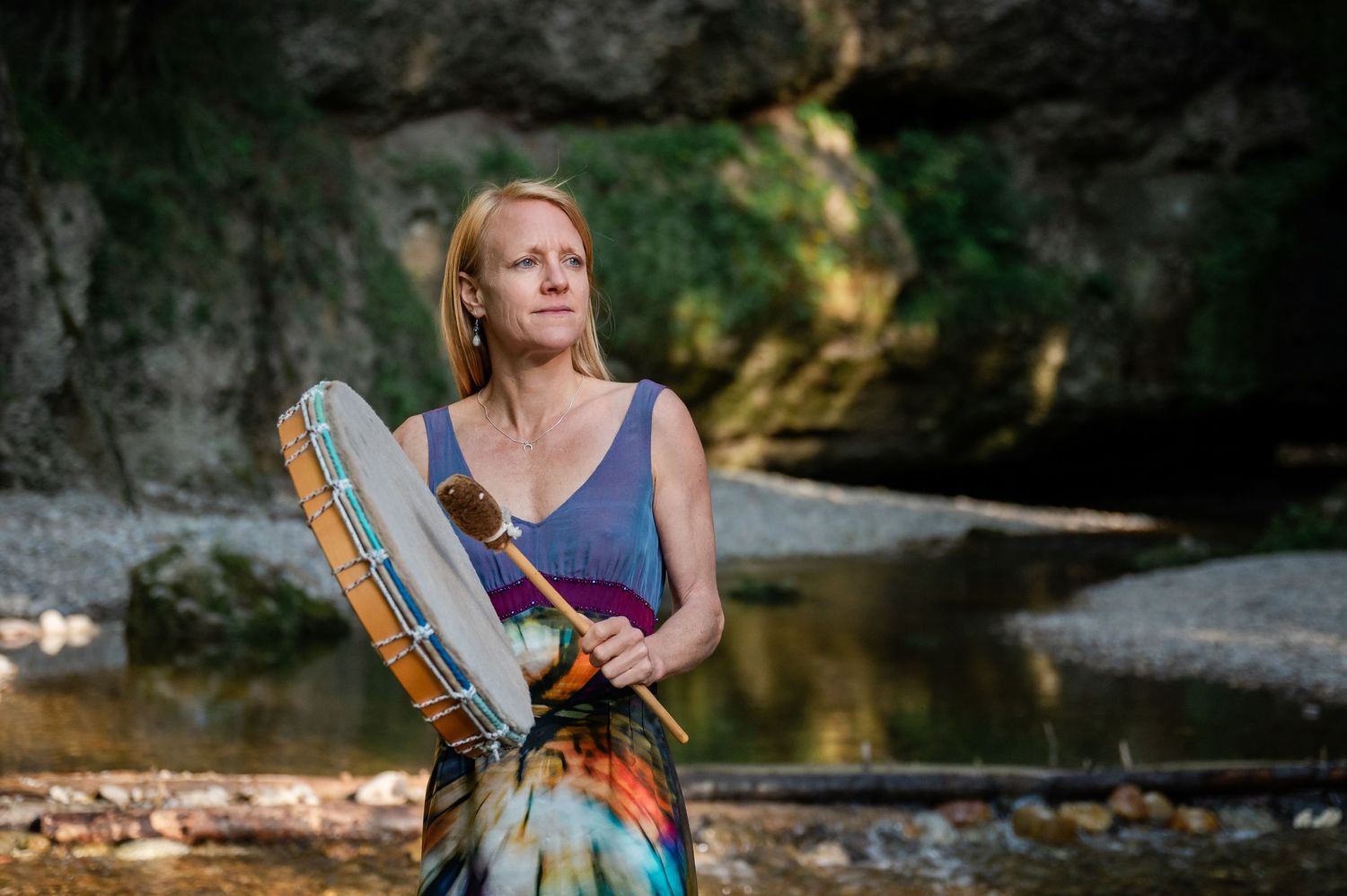 Frau mit schamanischer Trommel steht im langen Kleid vor Wasserfall – spirituelle Verbindung zur Natur im Abendlicht.