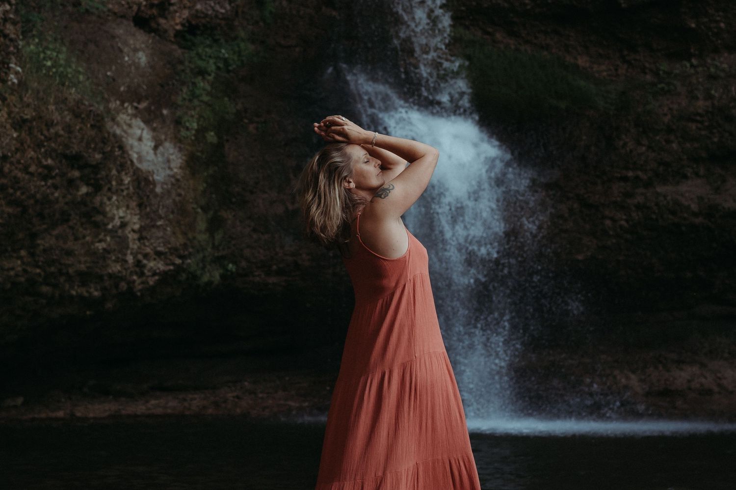 Frau im fliessenden Kleid posiert an einem Wasserfall – sinnliches Naturshooting mit Fokus auf Weiblichkeit und Freiheit.
