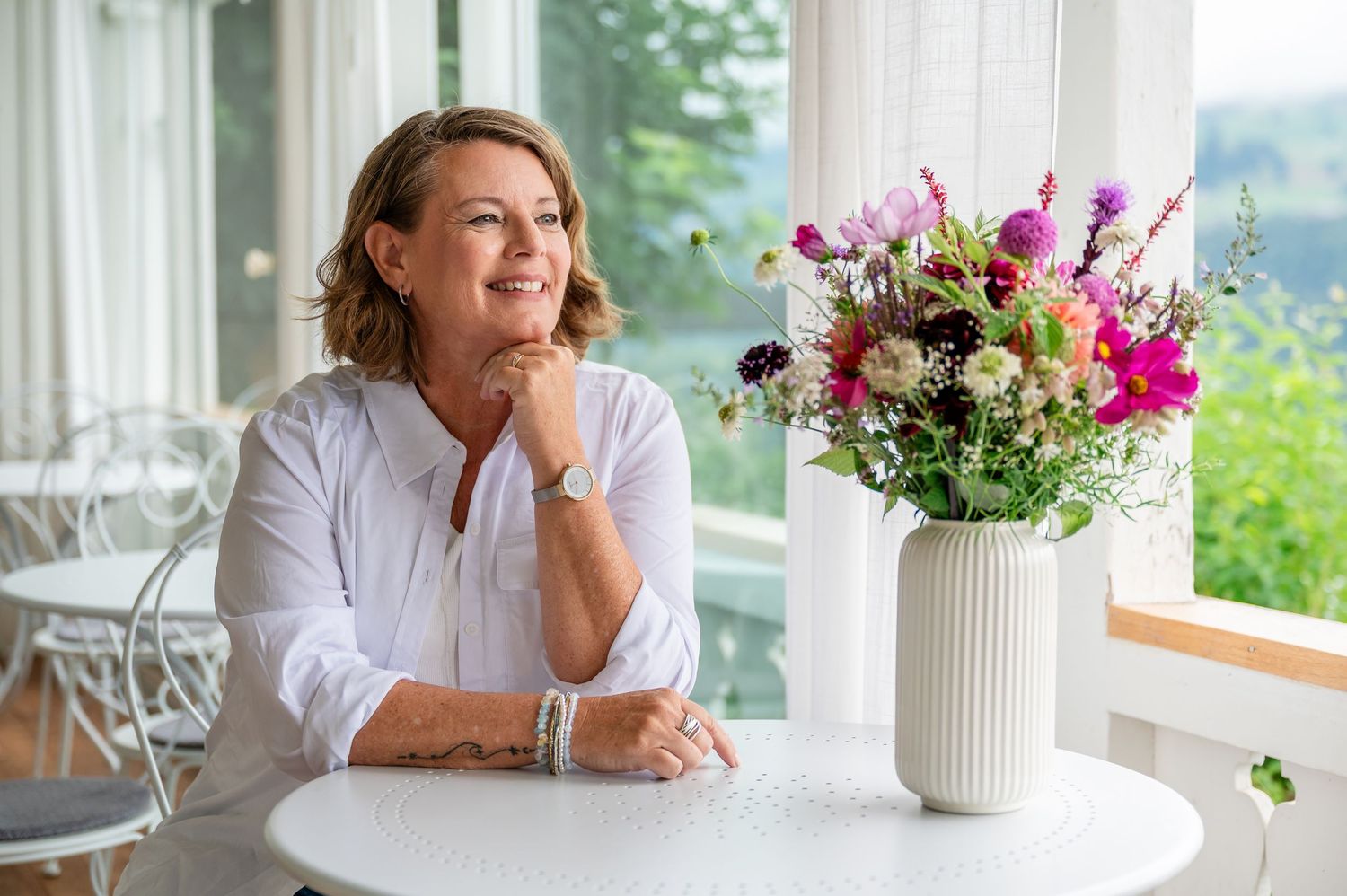 Frau mittleren Alters sitzt lächelnd am runden Tisch mit Sommerblumenstrauss, in heller Location mit Blick ins Grüne in Thun.