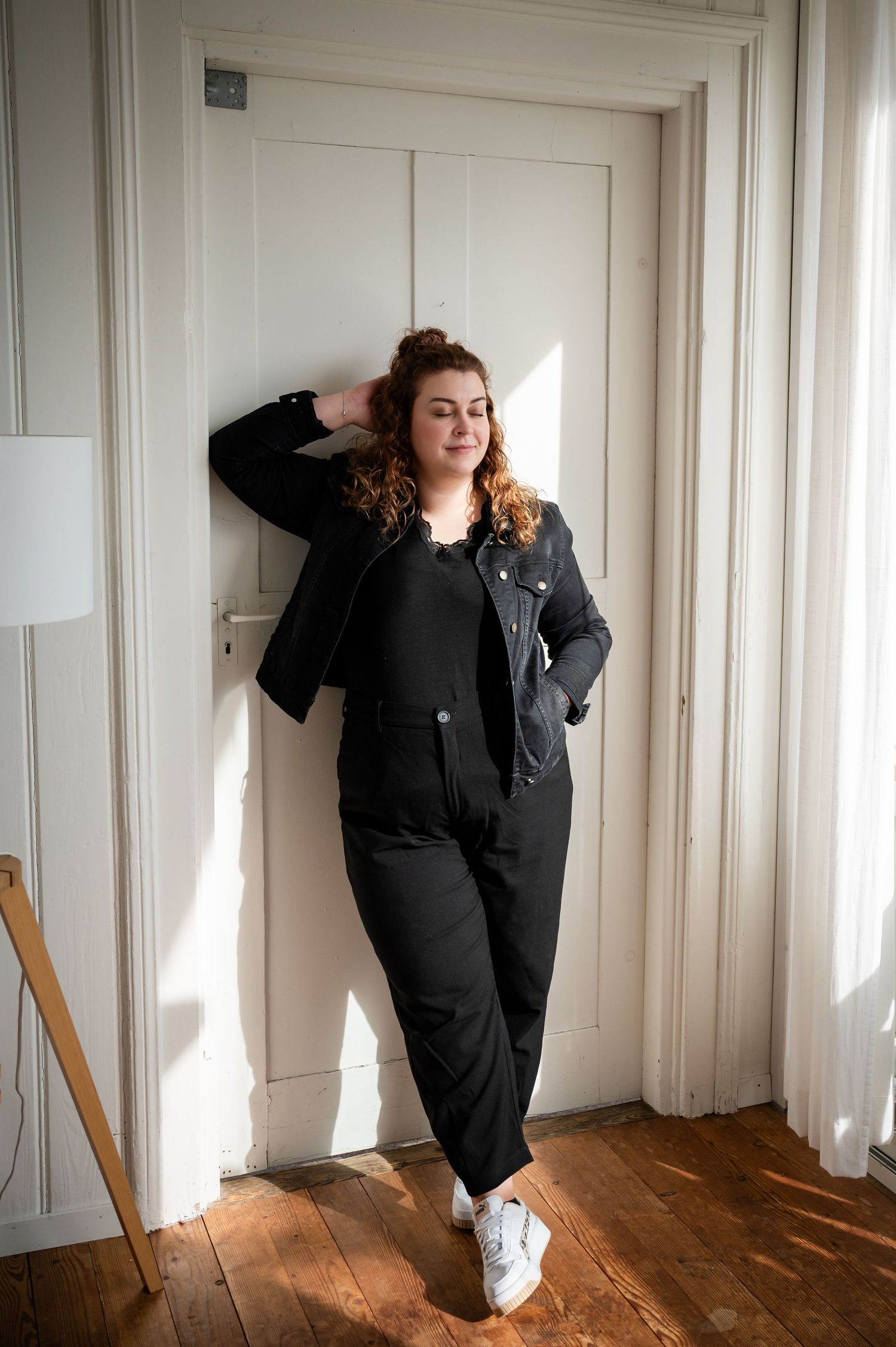 Junge Frau in schwarzem Outfit lehnt entspannt an Türrahmen, Sonnenlicht fällt durchs Fenster, selbstbewusste Ausstrahlung.