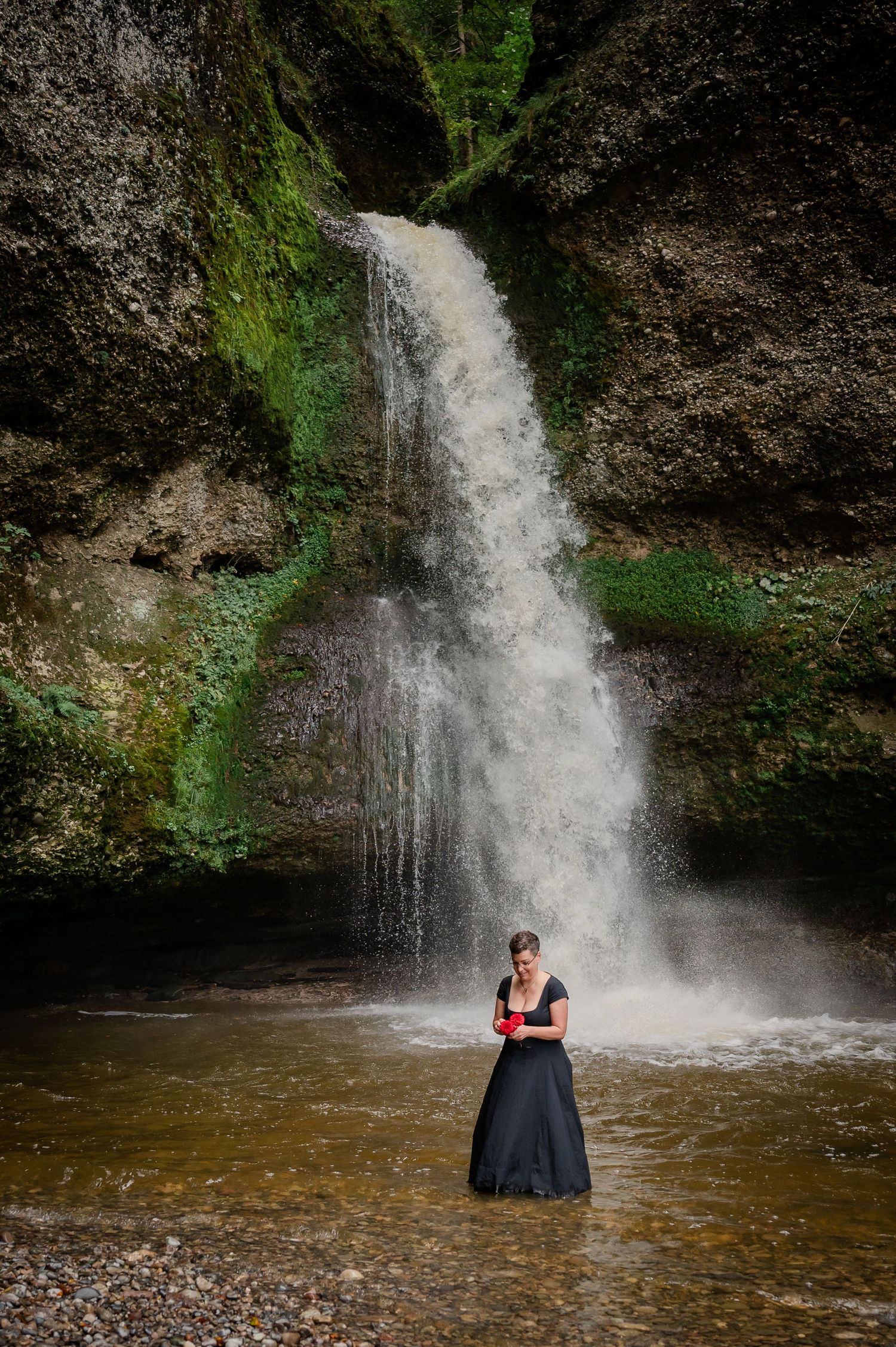 Figure in long dress stands in pool beneath dramatic waterfall in mossy canyon.