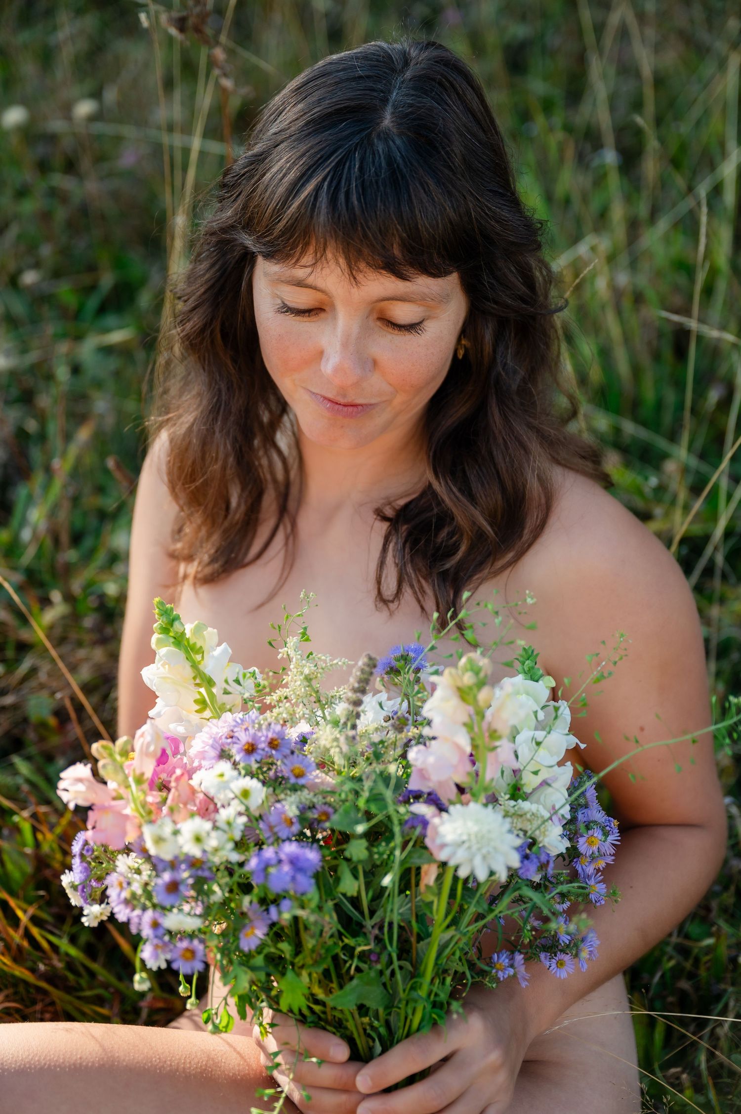 Natürliche Boudoir-Aufnahme einer Frau mit wildem Blumenstrauss, sanft lächelnd im warmen Licht auf einer Wiese sitzend.