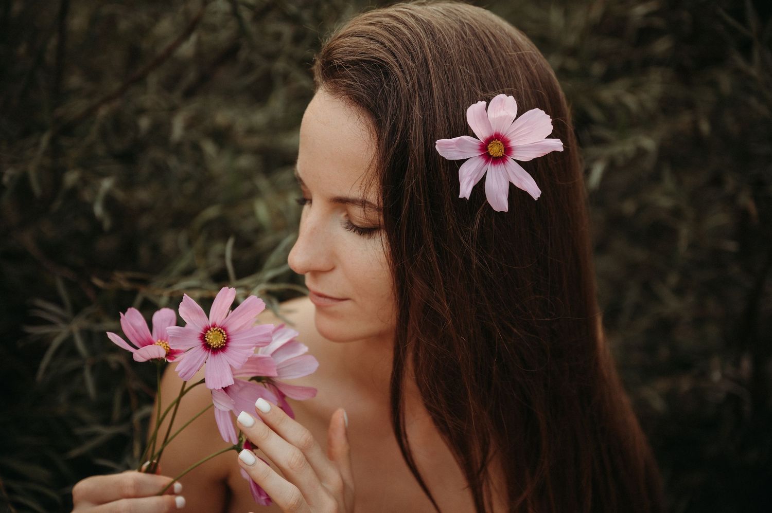 Frau mit langen Haaren und rosa Blumen im Haar hält zarte Blüten – natürliches Portrait mit sanfter, sinnlicher Ausstrahlung.