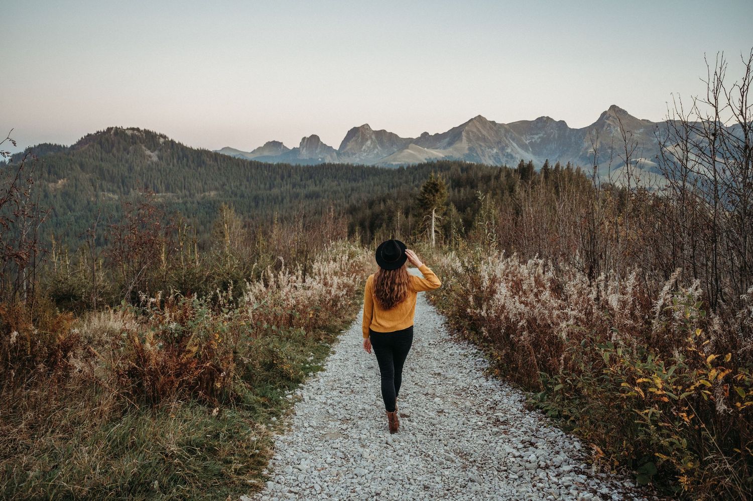 Frau mit Hut spaziert auf Kiesweg im Gantrischgebiet, umgeben von herbstlicher Natur und Bergpanorama – natürliches Outdoor-Porträt.
