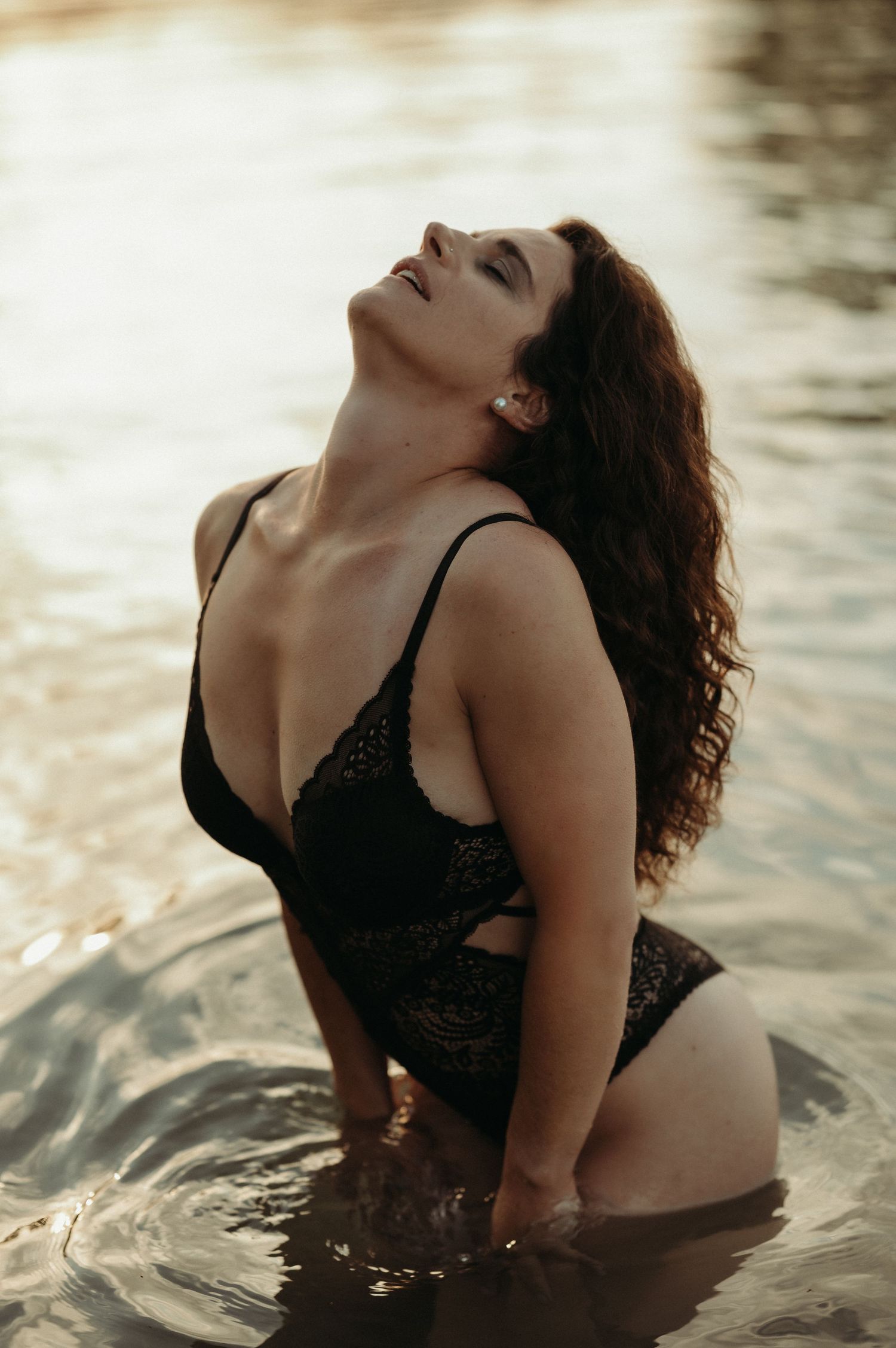 Sinnliches Boudoirshooting im Wasser bei Thun: Frau in schwarzer Spitze posiert selbstbewusst kniend im See im Abendlicht.