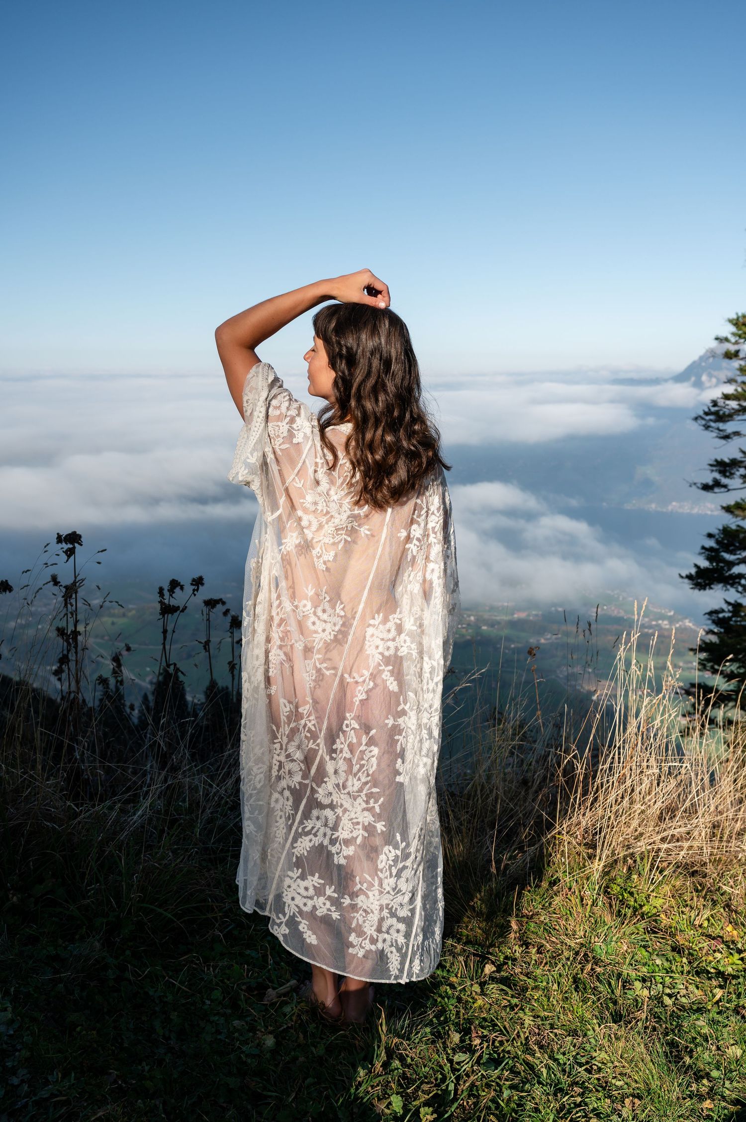 Frau in transparentem Spitzenkleid steht barfuß auf einer Wiese über den Wolken, umgeben von Himmel und Natur – Boudoir im Berner Oberland.