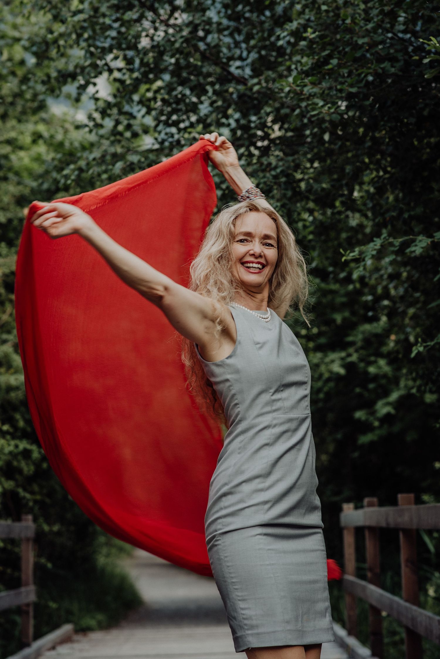 Strahlende Frau mit rotem Tuch und grauem Kleid in der Natur – lebendig, kraftvoll und voller Lebensfreude auf einem Holzsteg.