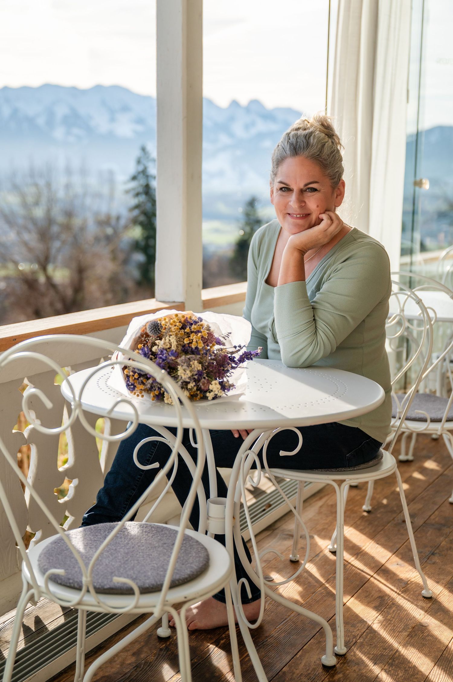 Frau mit grauem Dutt sitzt entspannt auf einer sonnigen Veranda mit Alpenblick, neben ihr ein Strauss Trockenblumen – Selbstliebe-Shooting