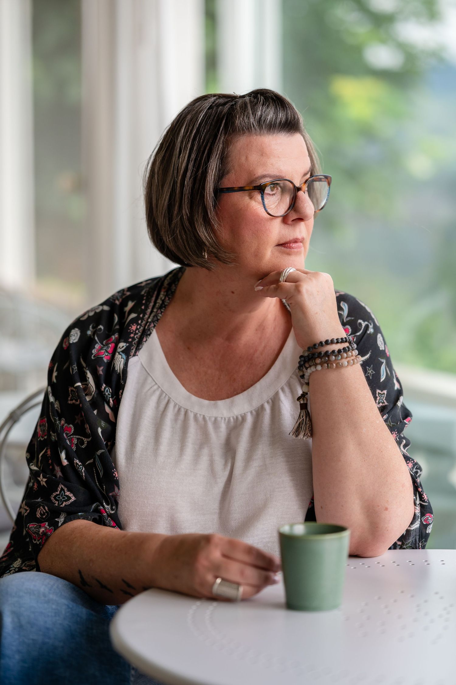 Person in casual attire sitting thoughtfully by a window with a teal coffee mug.