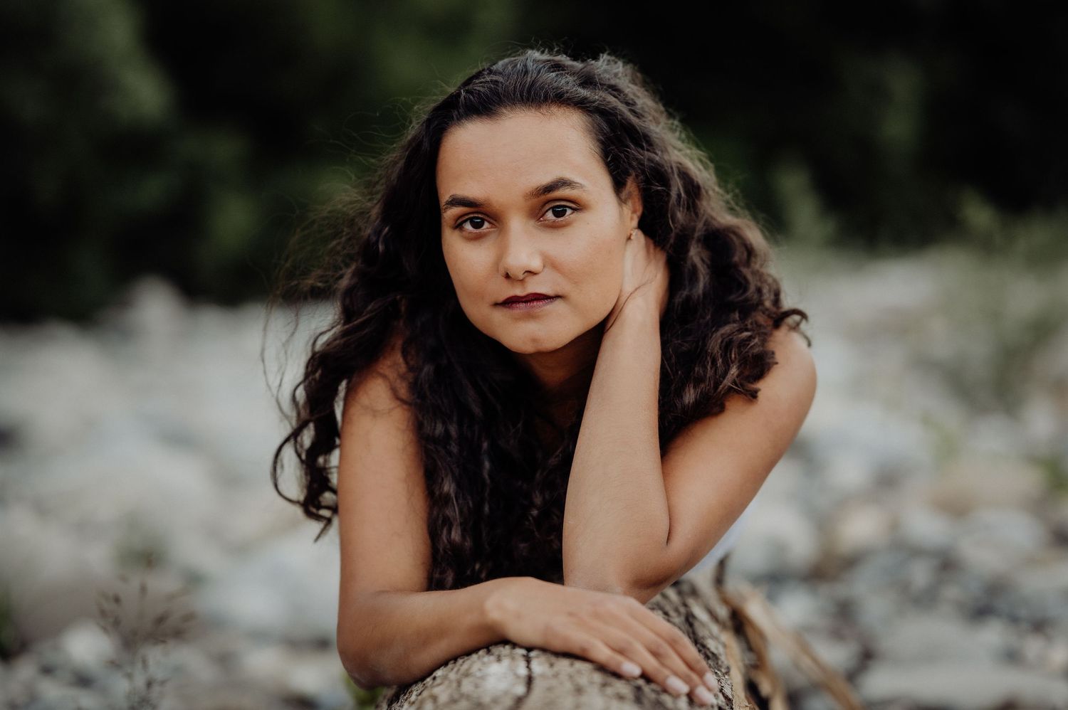 Portrait einer Frau mit langen Locken, die auf einem Baumstamm liegt und direkt in die Kamera blickt. Aufnahme am Ufer bei Thun.