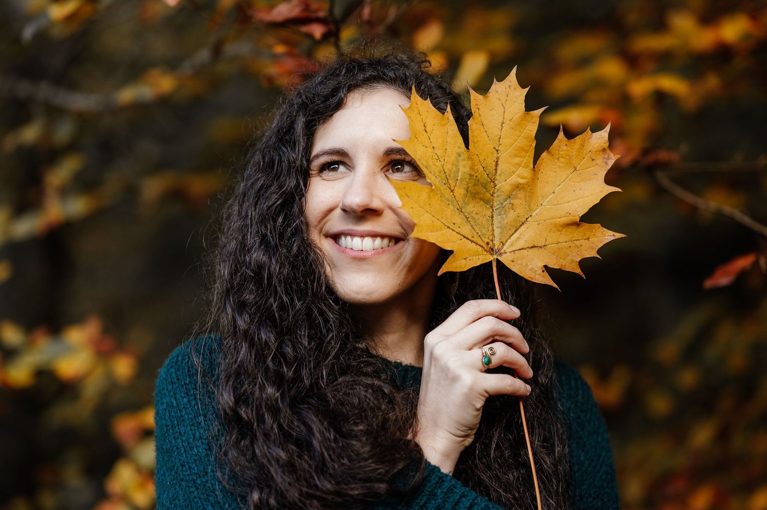 Person with curly hair holds autumn maple leaf while smiling outdoors.