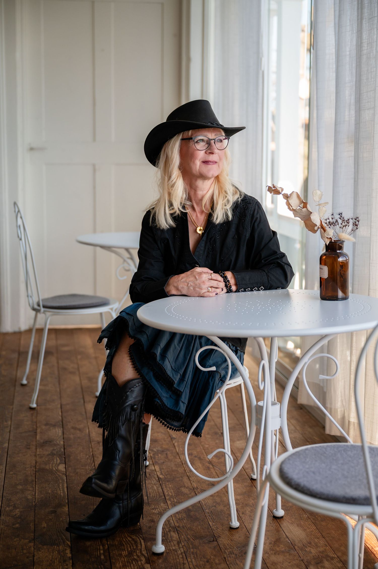 Stilvolle Frau mit Cowboyhut und Stiefeln sitzt an einem runden Tisch im Sonnenlicht – selbstbewusst, entspannt und authentisch.