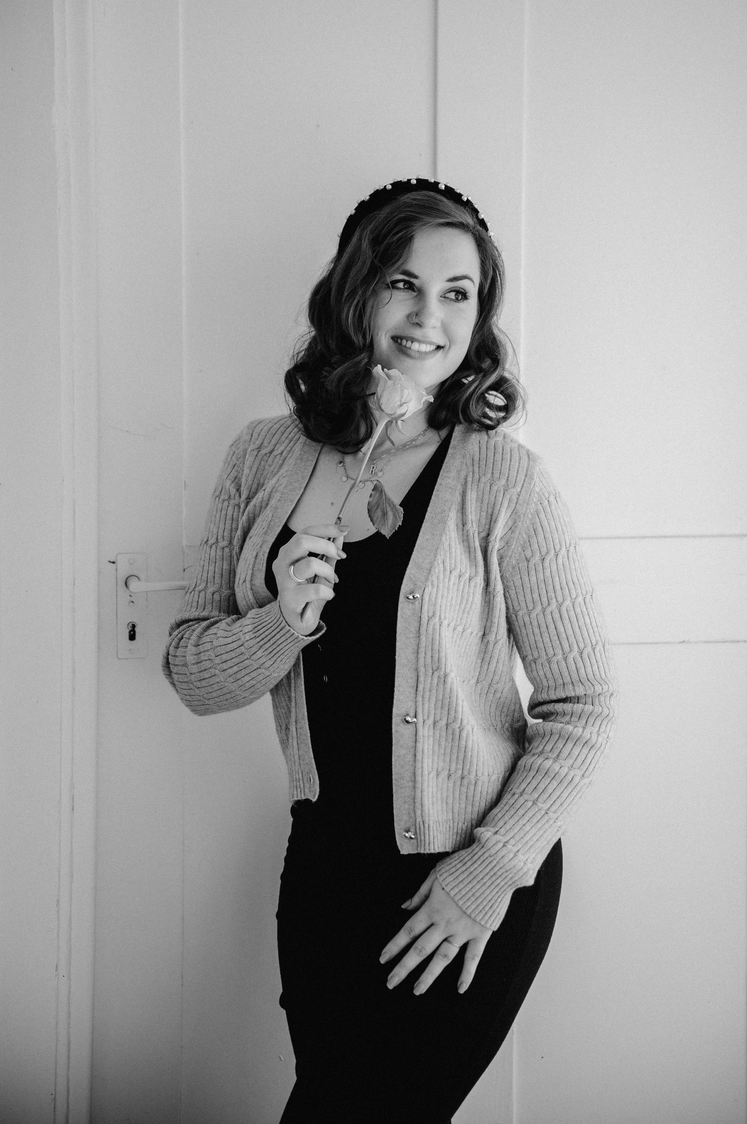 Frau mit Locken, Haarreif und Rose in der Hand lehnt lächelnd an einer Tür, in elegantem Schwarz-Weiß-Porträt – Selbstliebe-Shooting.