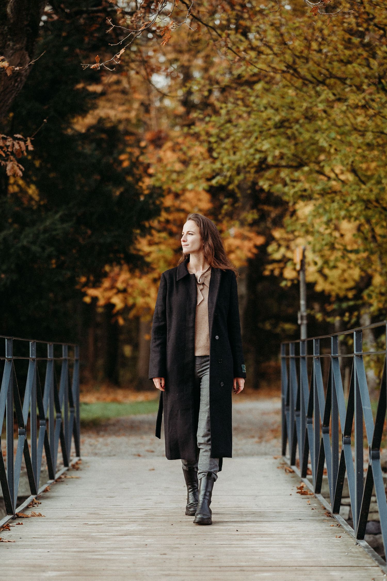 Frau im langen Mantel spaziert über eine Brücke im herbstlich gefärbten Wald – stimmungsvolles Outdoor-Herbstshooting.