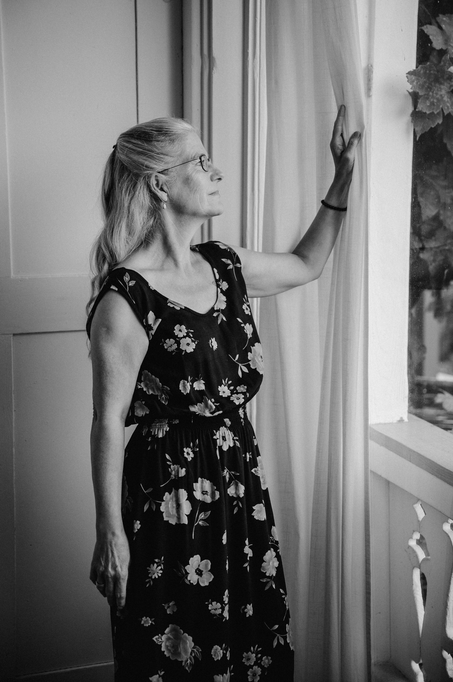Schwarzweissporträt einer älteren Frau mit Brille im geblümten Kleid, die nachdenklich aus einem Fenster schaut und den Vorhang hält.